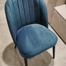 MeblePasternak - Turkusowe krzesło tapicerowane z pionowymi przeszyciami na oparciu, z ciemnymi nogami, na tle beżowej sofy i płytek podłogowych.