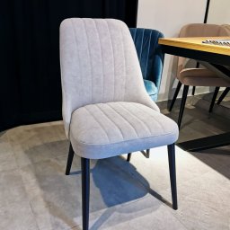 MeblePasternak - Tapicerowane krzesło w odcieniach szarości z pionowymi przeszyciami na siedzisku i oparciu, czarne nogi, w tle inne krzesła i fragment stołu.