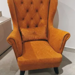 Pomarańczowy fotel uszak z guzikami i poduszką, stojący na szarej podłodze z płytek, z widocznym fragmentem białej komody w tle.