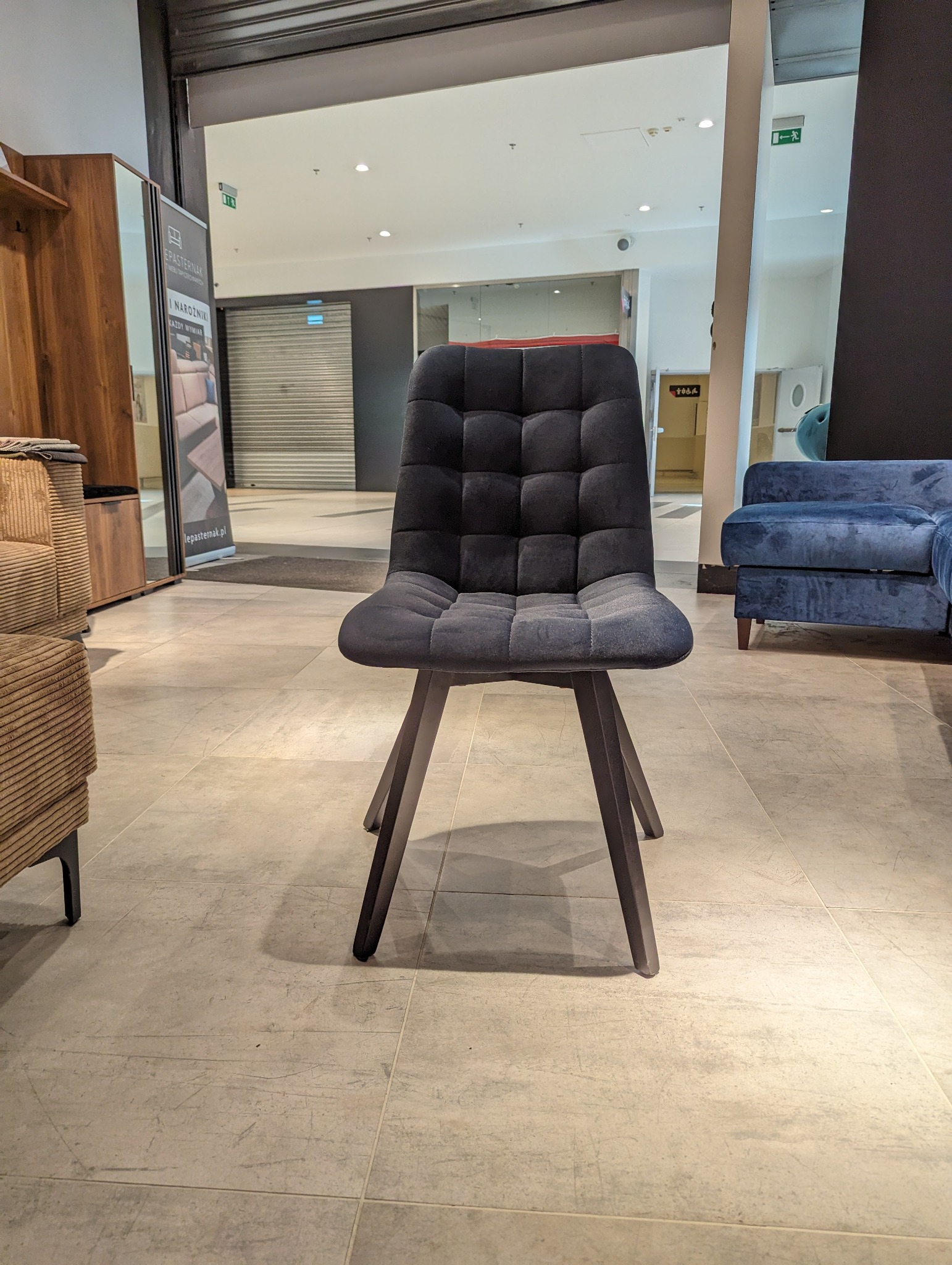 Ciemnoszare krzesło tapicerowane z kwadratowymi przeszyciami, ustawione na tle salonu meblowego z widocznymi innymi meblami i zamkniętymi roletami sklepowymi.