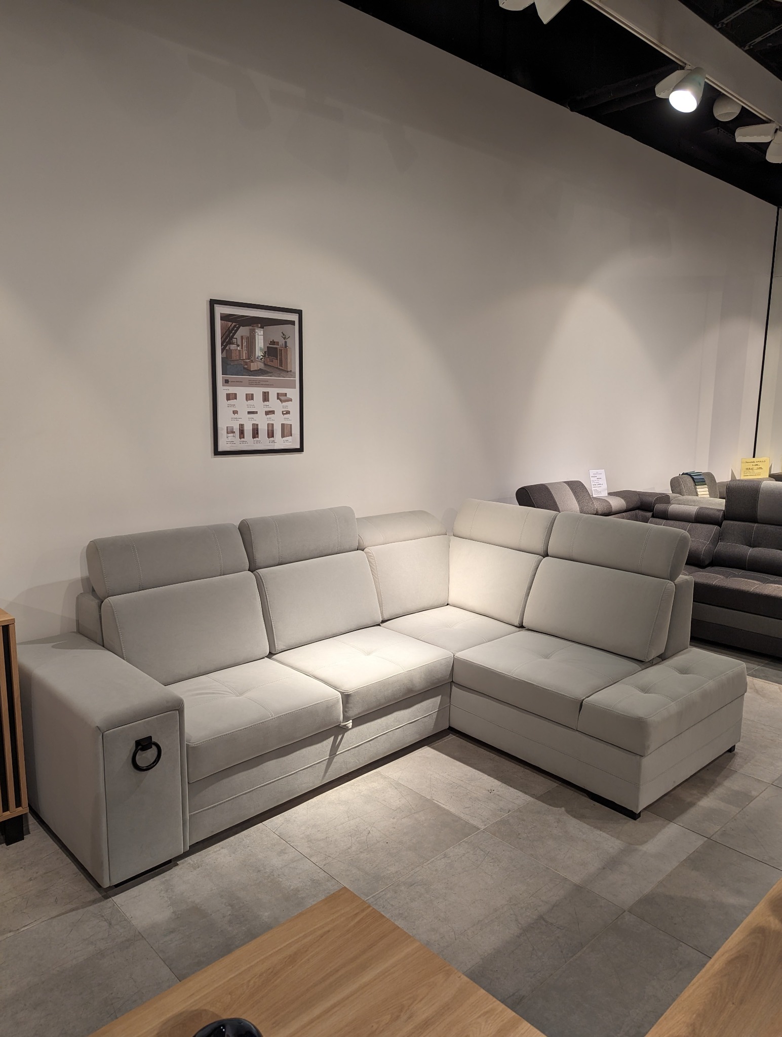 Jasnoszara, modułowa sofa narożna z regulowanymi zagłówkami i metalowym uchwytem w kształcie pierścienia na boku, prezentowana w salonie meblowym.