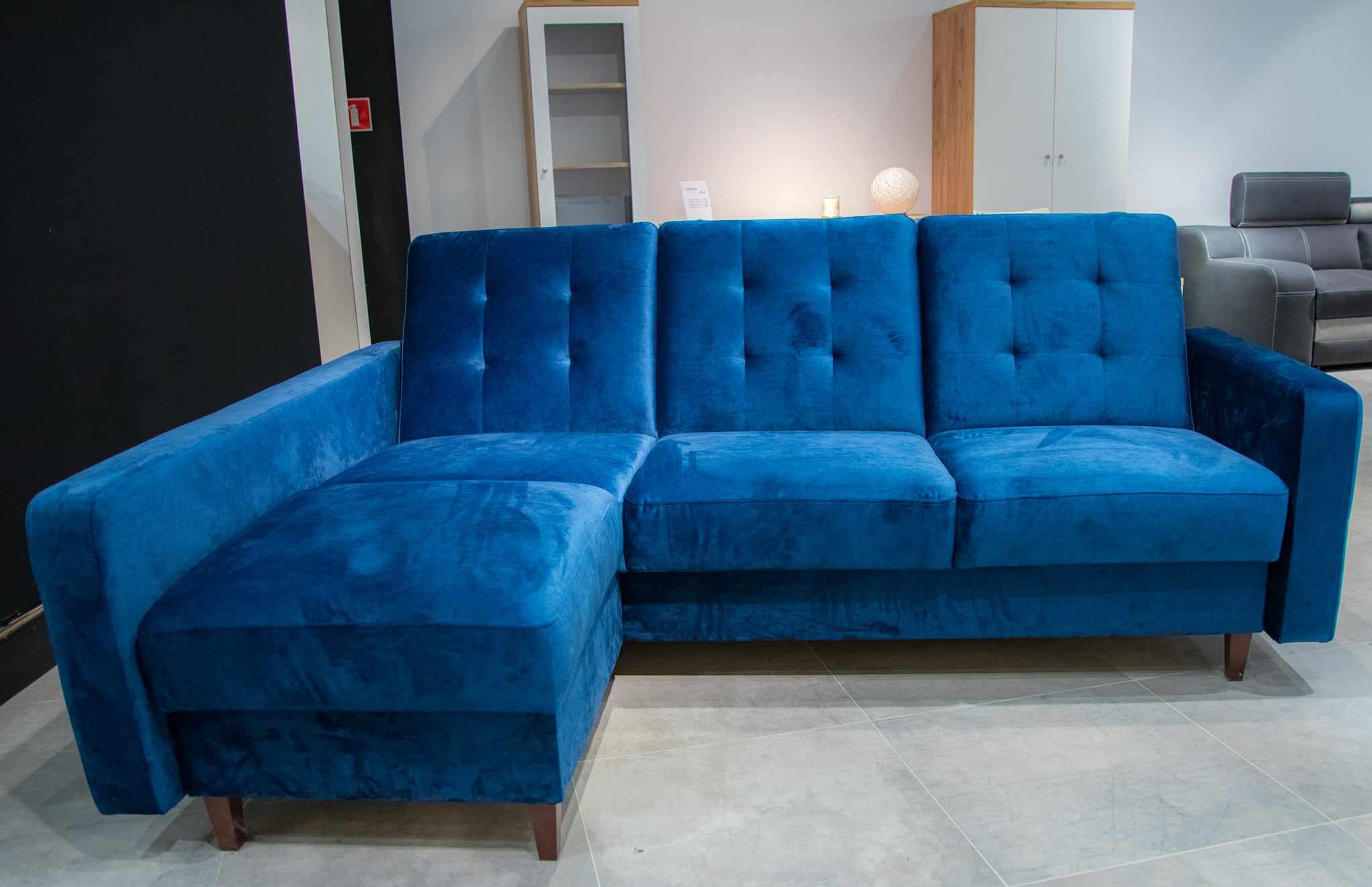 Rozłożysta, nowoczesna sofa narożna obita niebieskim welurem z niskimi, drewnianymi nóżkami, na tle jasnej ściany i szafek.