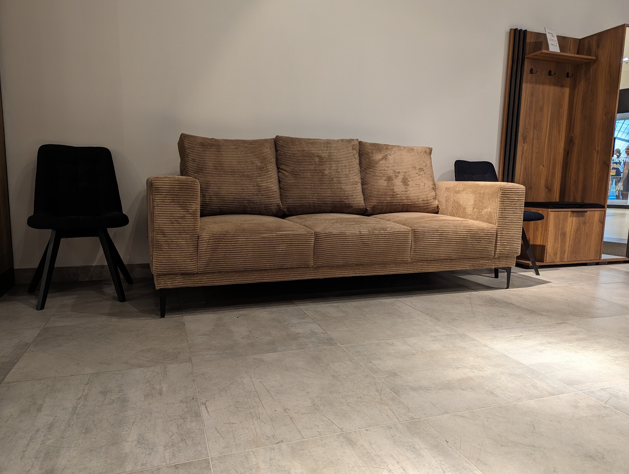 Trzyosobowa sofa w prążkowanej, beżowej tapicerce, z trzema poduszkami, na tle jasnej ściany i podłogi z płytek, obok stoi czarne krzesło i drewniany wieszak z szafką.
