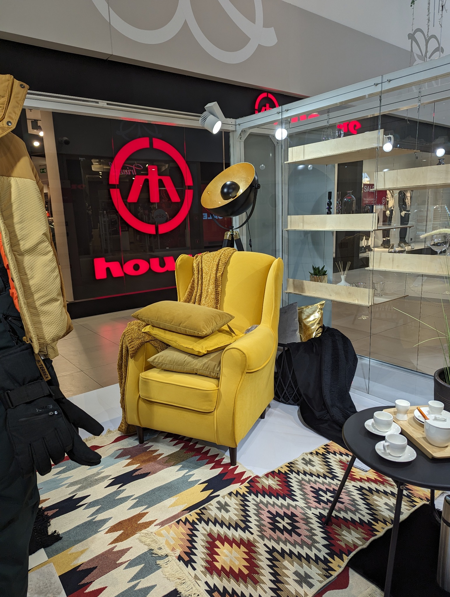 Żółty fotel z poduszkami i pledem na wzorzystym dywanie, obok stolik z filiżankami, w tle ekspozycja sklepu odzieżowego.