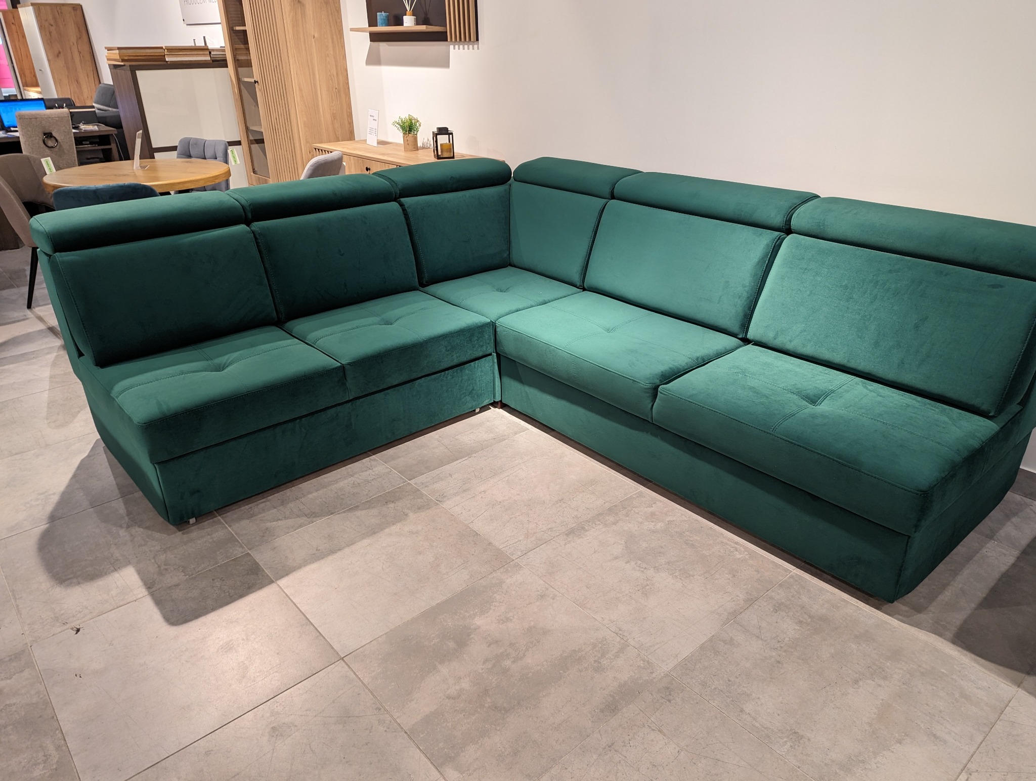 Zielony, narożny zestaw wypoczynkowy z aksamitną tapicerką, widoczny na tle jasnego wnętrza salonu meblowego.