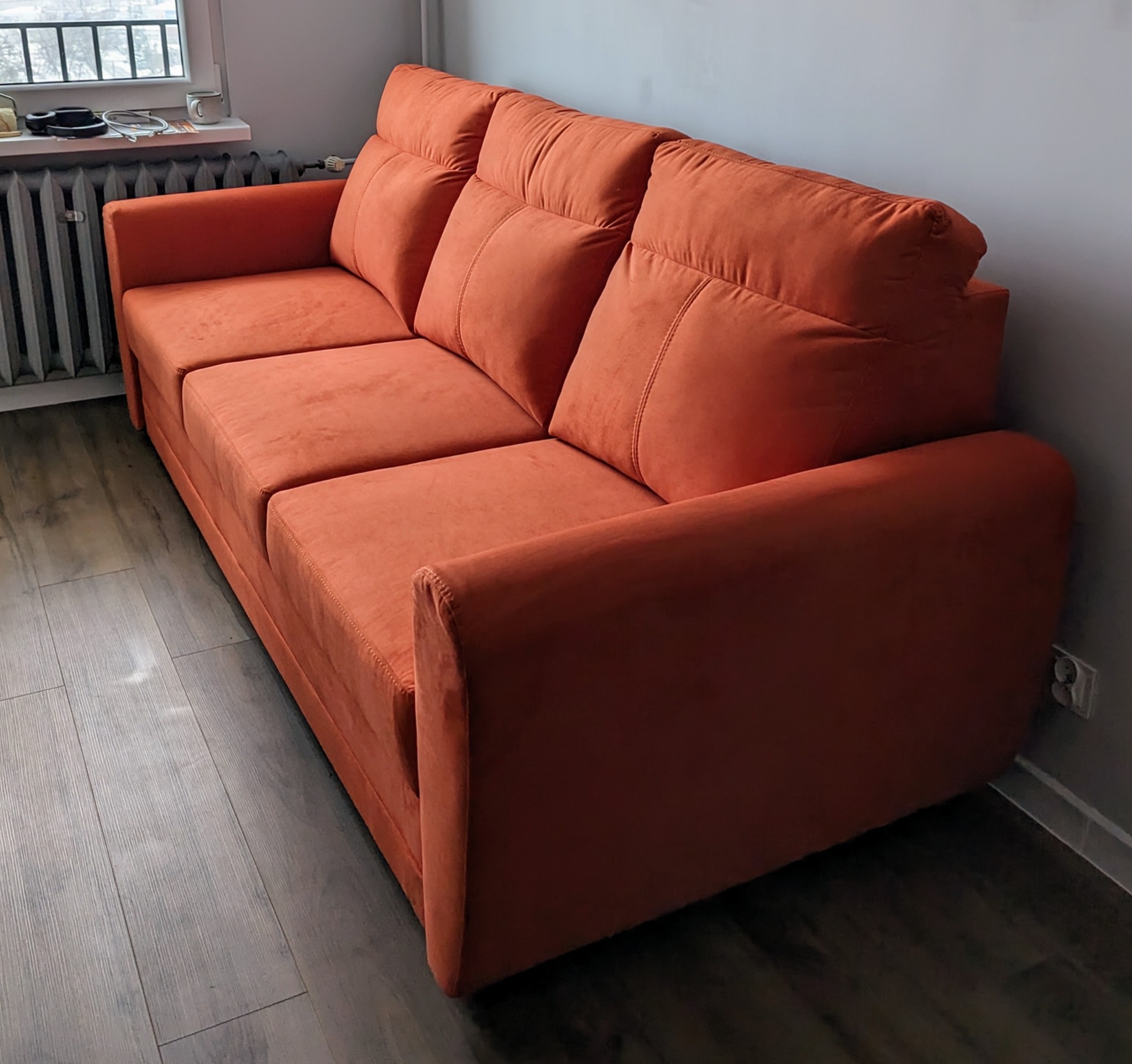 Pomarańczowa, tapicerowana sofa z trzema poduszkami na drewnianej podłodze, widok z boku.