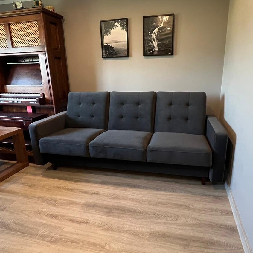 Szara, trzyosobowa sofa z guzikami na oparciu, ustawiona w salonie z drewnianą podłogą i starym pianinem z tyłu, na ścianie wiszą dwa czarno-białe obrazy.