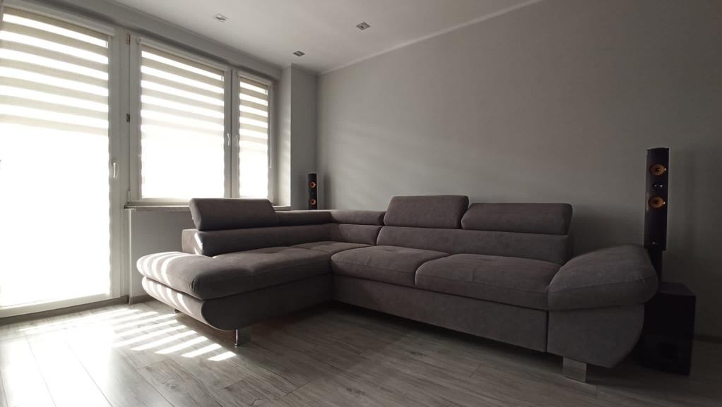 Szary narożnik z regulowanymi zagłówkami w jasnym salonie, obok okna z żaluzjami dzień-noc i wysokich głośników.