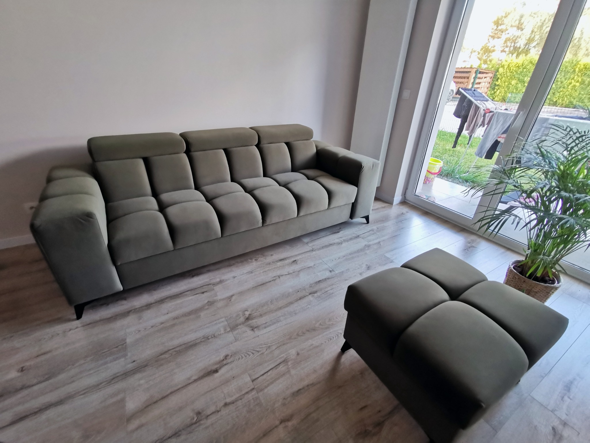 Nowoczesna, oliwkowa sofa tapicerowana z pufą w minimalistycznym salonie, widok z boku, drewnopodobna podłoga, roślina doniczkowa przy oknie.