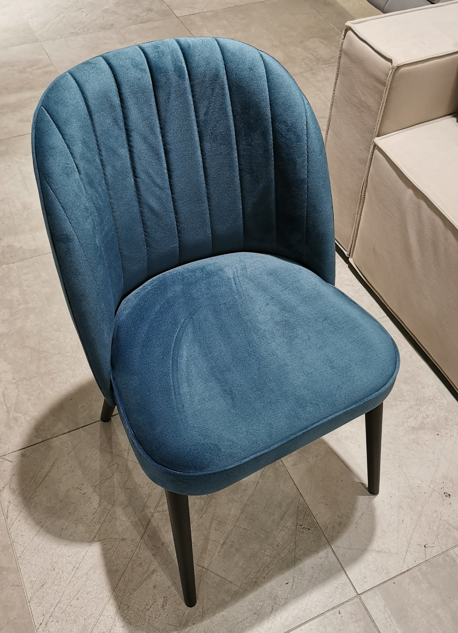 Turkusowe krzesło tapicerowane z pionowymi przeszyciami na oparciu, z ciemnymi nogami, na tle beżowej sofy i płytek podłogowych.