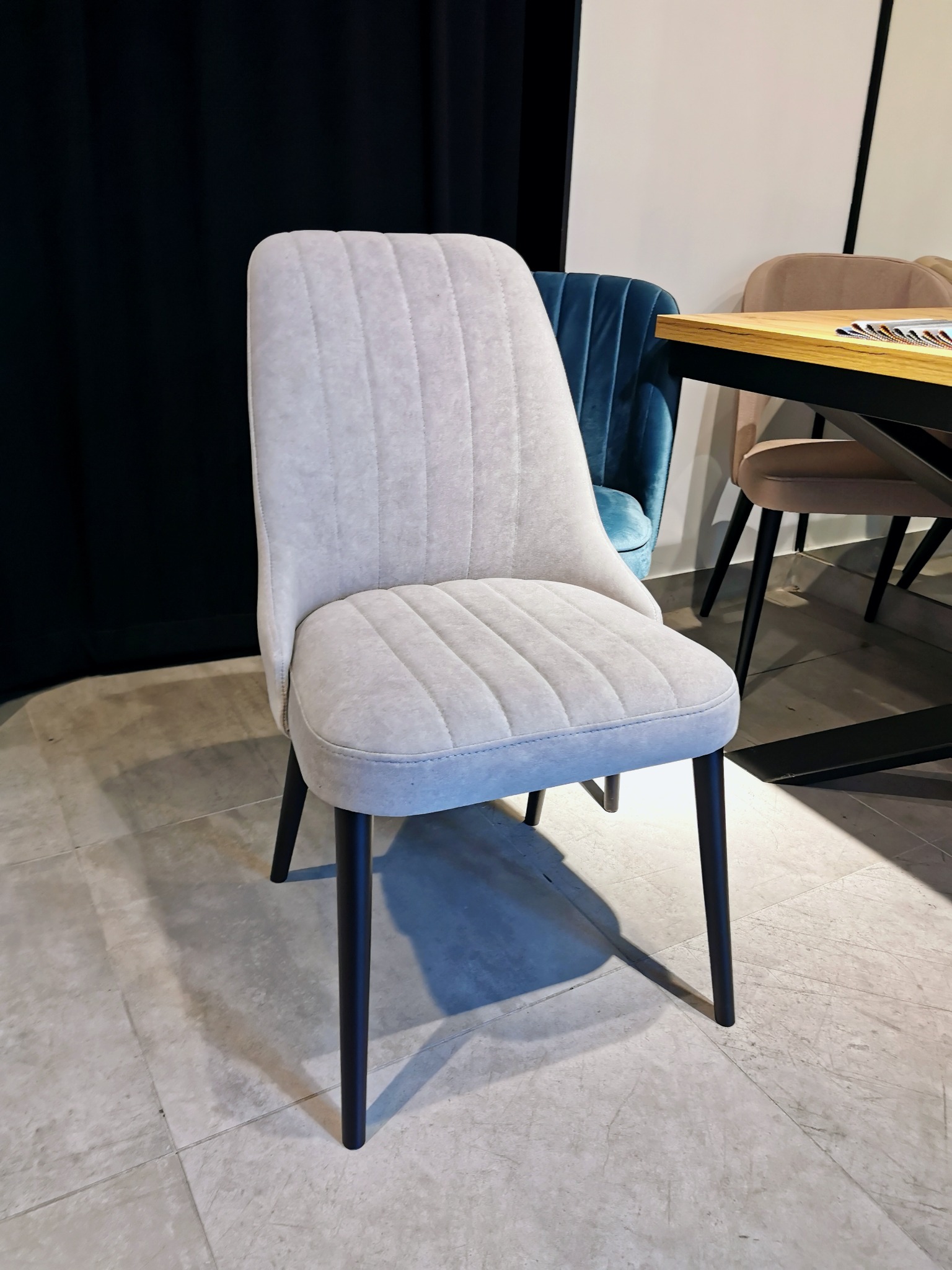 Tapicerowane krzesło w odcieniach szarości z pionowymi przeszyciami na siedzisku i oparciu, czarne nogi, w tle inne krzesła i fragment stołu.