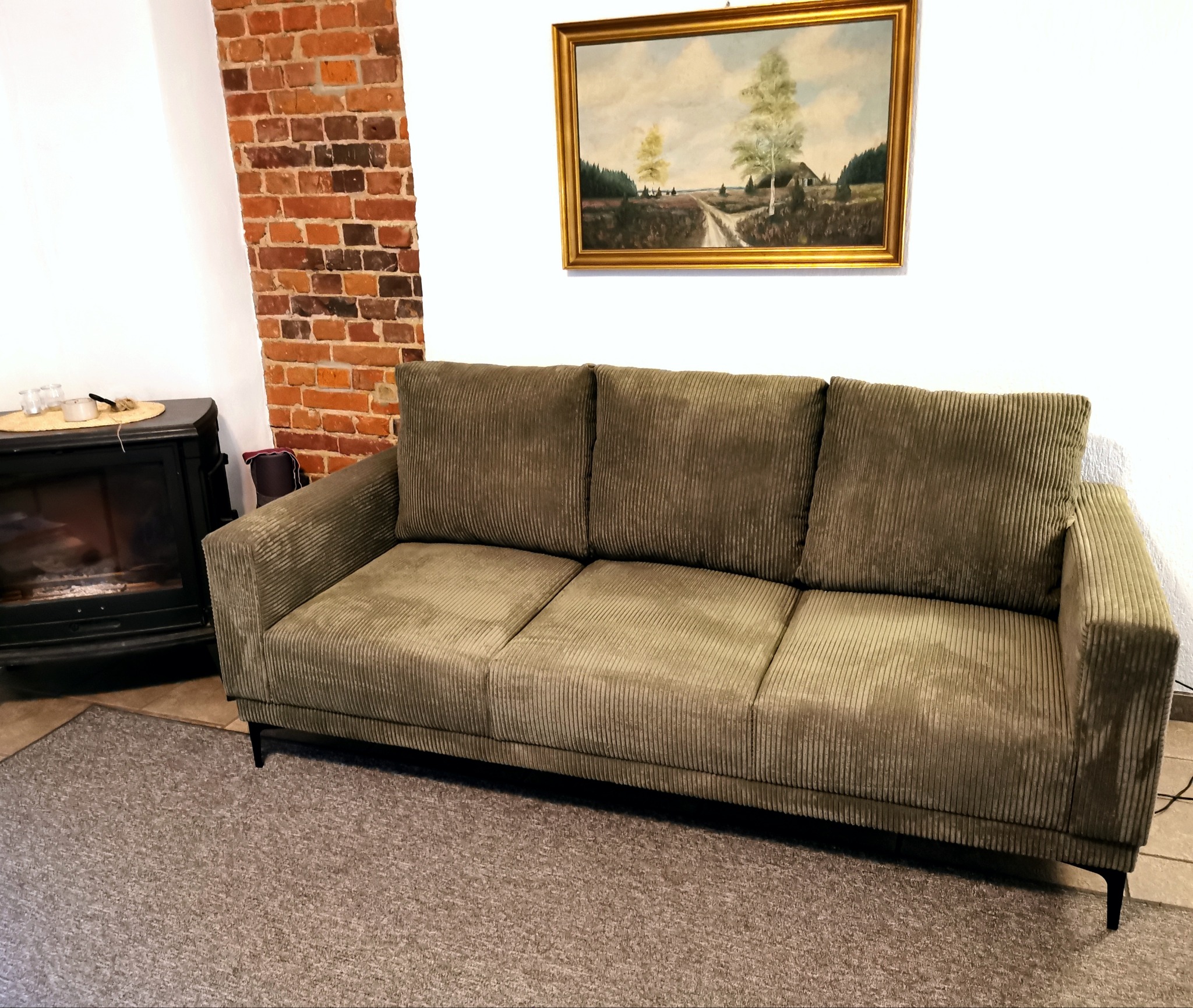 Trzyosobowa sofa tapicerowana sztruksem w odcieniach oliwki, na tle białej ściany z obrazem i fragmentem ściany z czerwonej cegły, obok kominek.