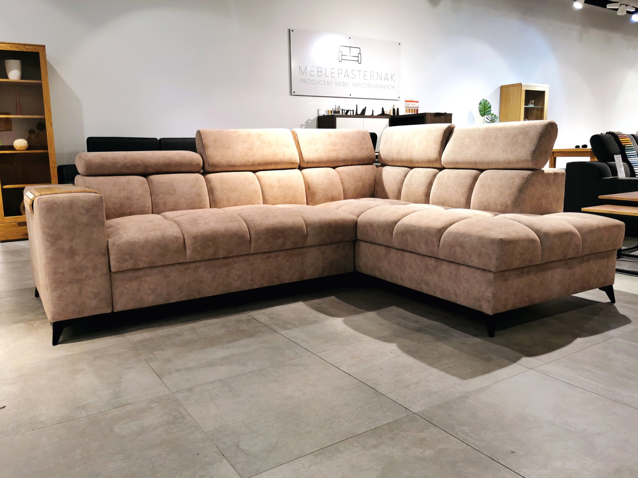 Narożnik tapicerowany w odcieniach beżu z regulowanymi zagłówkami, prezentowany w salonie meblowym z logo producenta widocznym w tle.