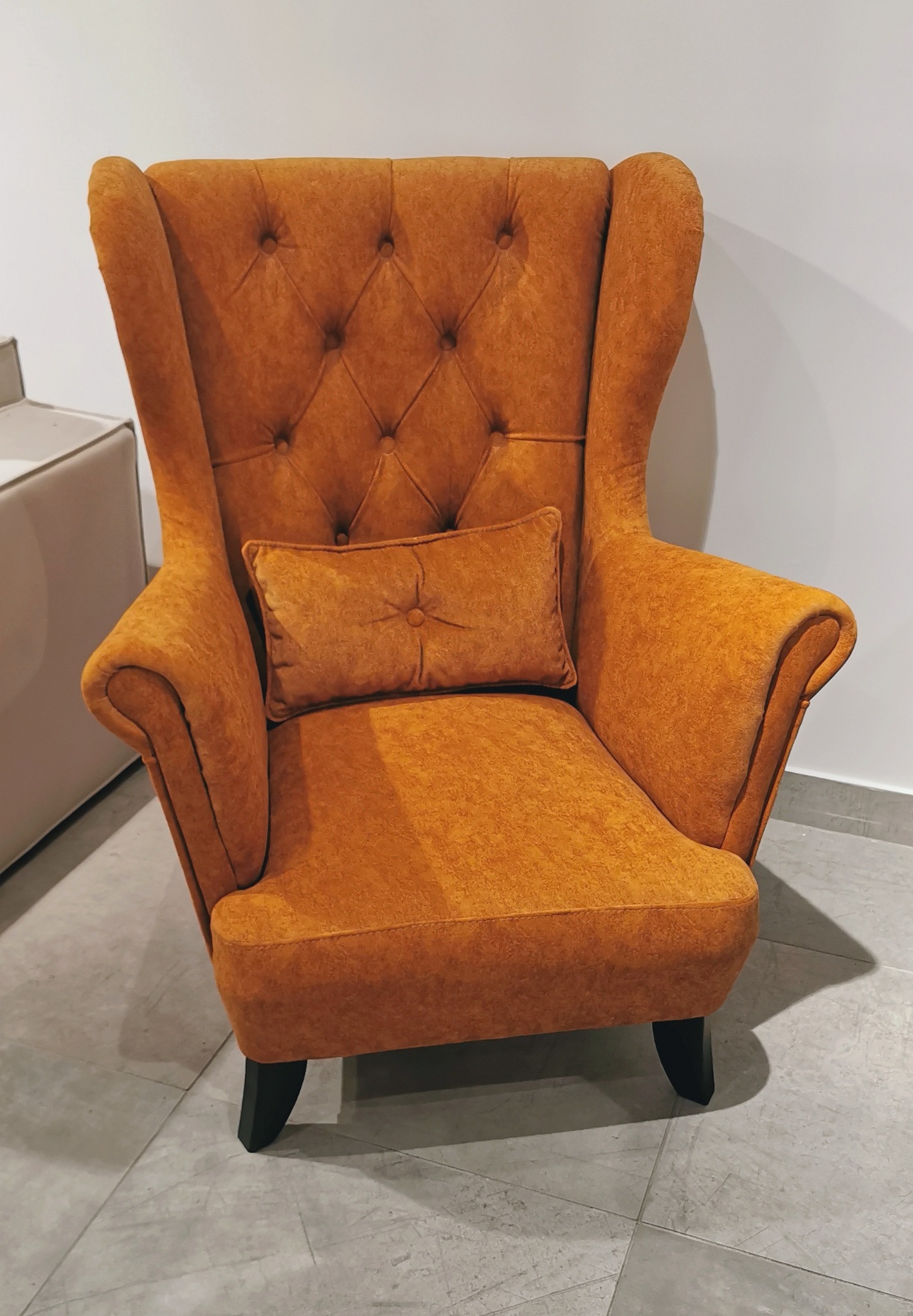 Pomarańczowy fotel uszak z guzikami i poduszką, stojący na szarej podłodze z płytek, z widocznym fragmentem białej komody w tle.