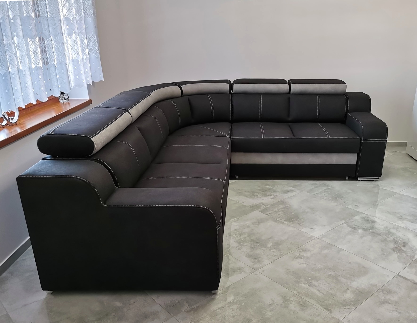 Nowoczesny narożnik tapicerowany w odcieniach szarości i czerni, z dekoracyjnymi przeszyciami, ustawiony w jasnym pomieszczeniu z oknem i marmurową podłogą.