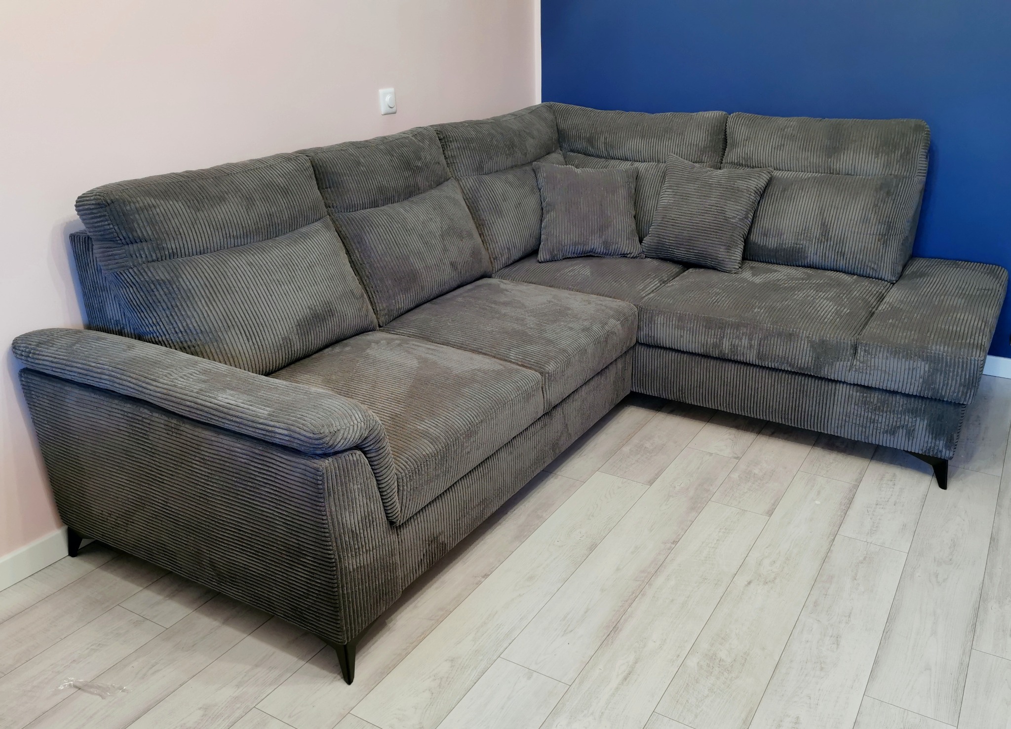 Szary narożnik z tkaniny w prążki z poduszkami, na drewnianej podłodze, na tle ściany w kolorze różowym i niebieskim.