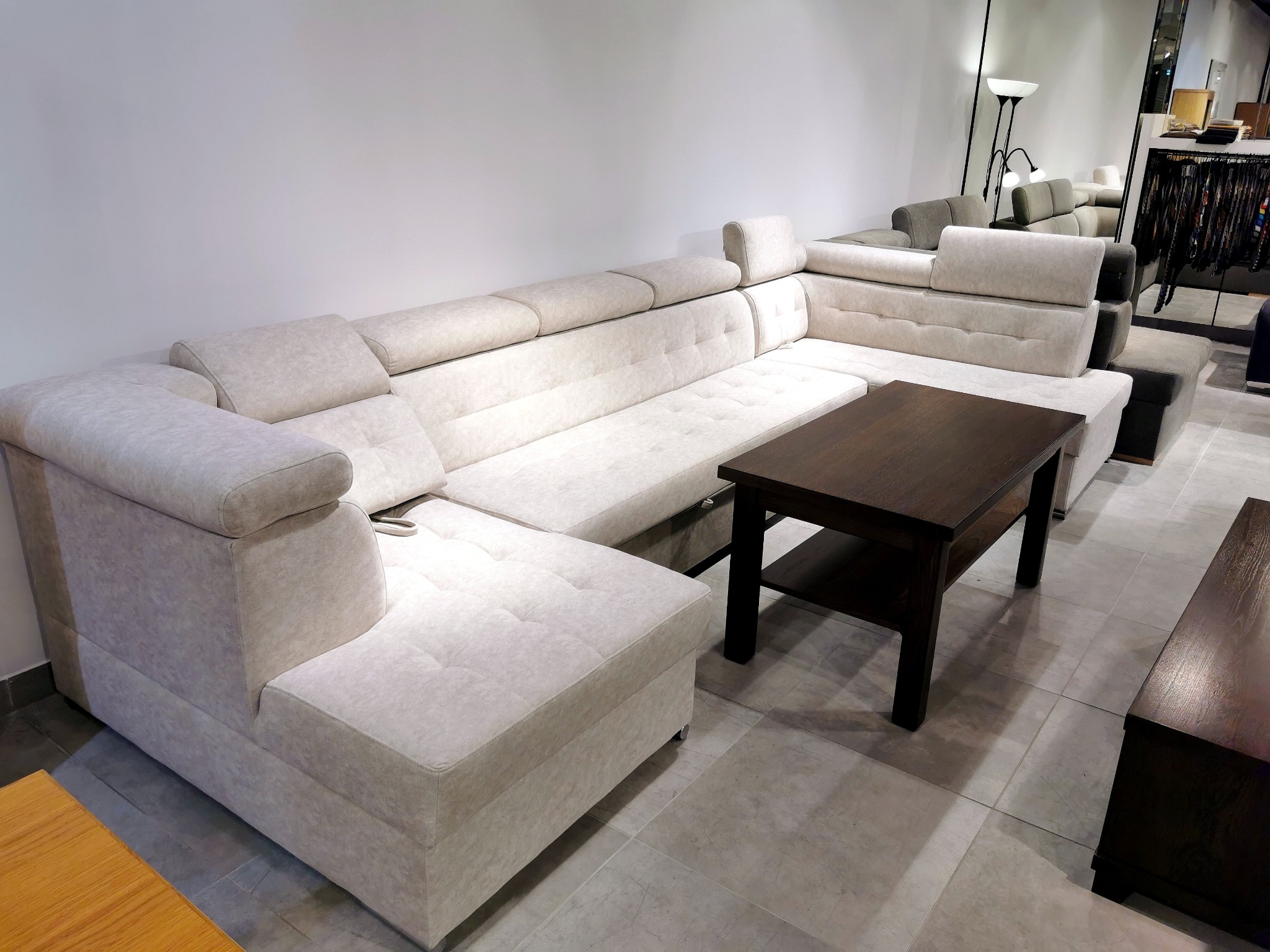 Jasny narożnik tapicerowany z regulowanymi zagłówkami i pikowanymi siedziskami, uzupełniony ciemnym stolikiem kawowym z półką, na tle jasnych ścian i podłogi w salonie meblowym.