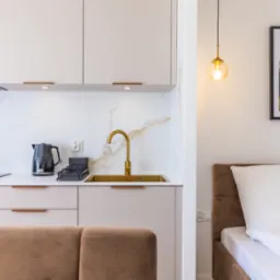 Nowoczesna kuchnia z elementami sypialni: złota armatura, białe szafki, czajnik elektryczny i łóżko z brązowym zagłówkiem w tle.
