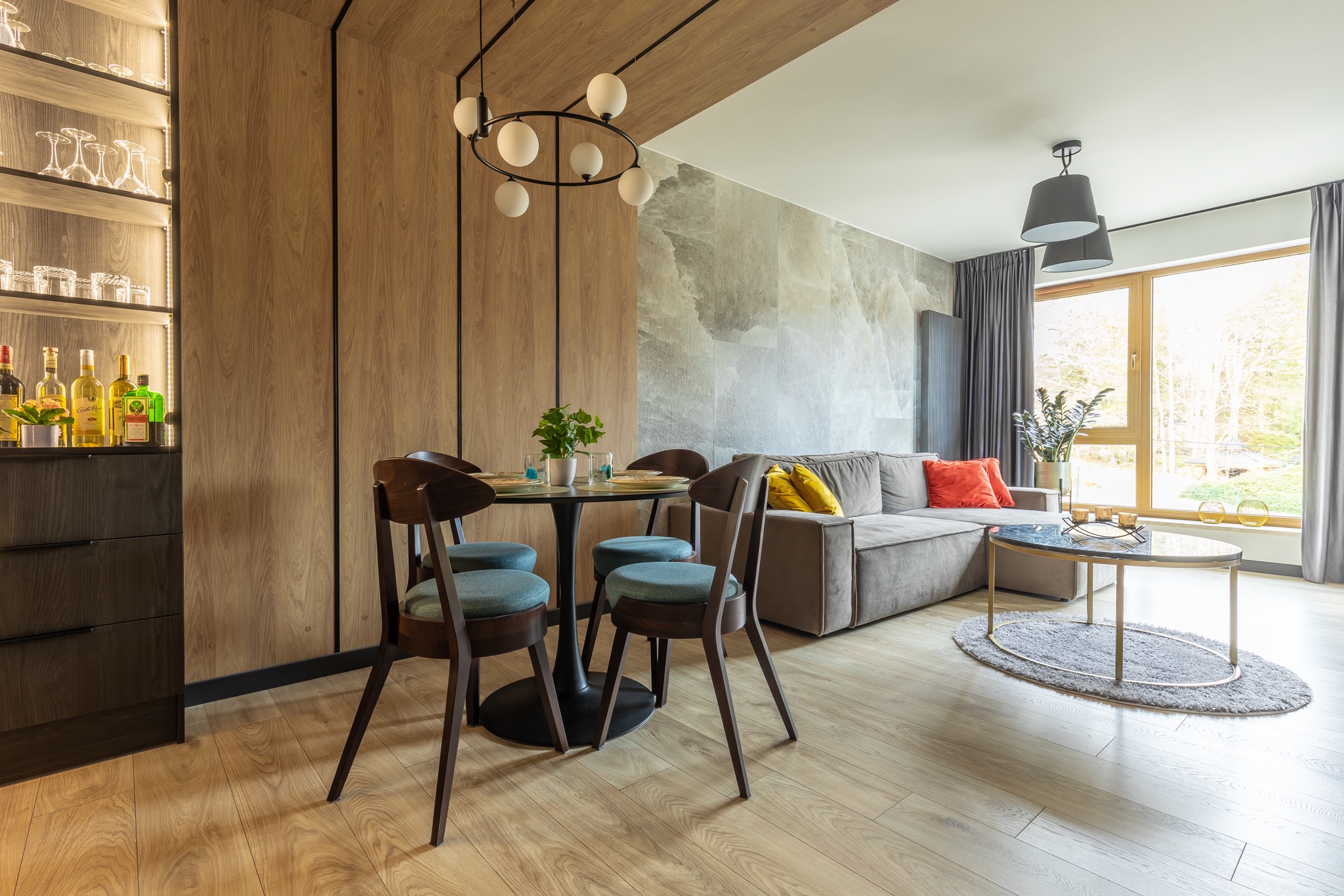 Jasne wnętrze mieszkania z drewnianą zabudową, okrągłym stołem jadalnym z czterema krzesłami, szarą sofą i stolikiem kawowym na tle okna.