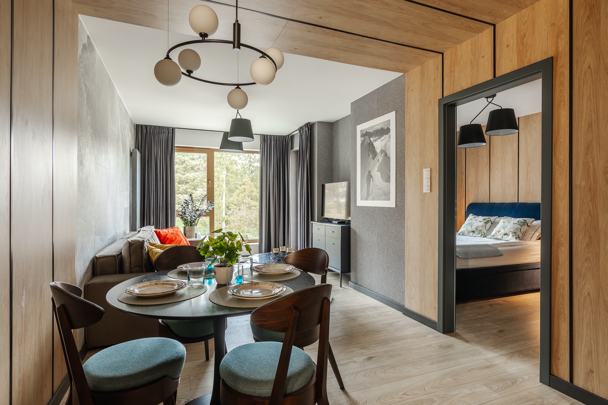 Nowoczesne wnętrze mieszkania z drewnianymi panelami ściennymi, okrągłym stołem jadalnym z turkusowymi krzesłami i widokiem na salon oraz sypialnię przez otwarte drzwi.