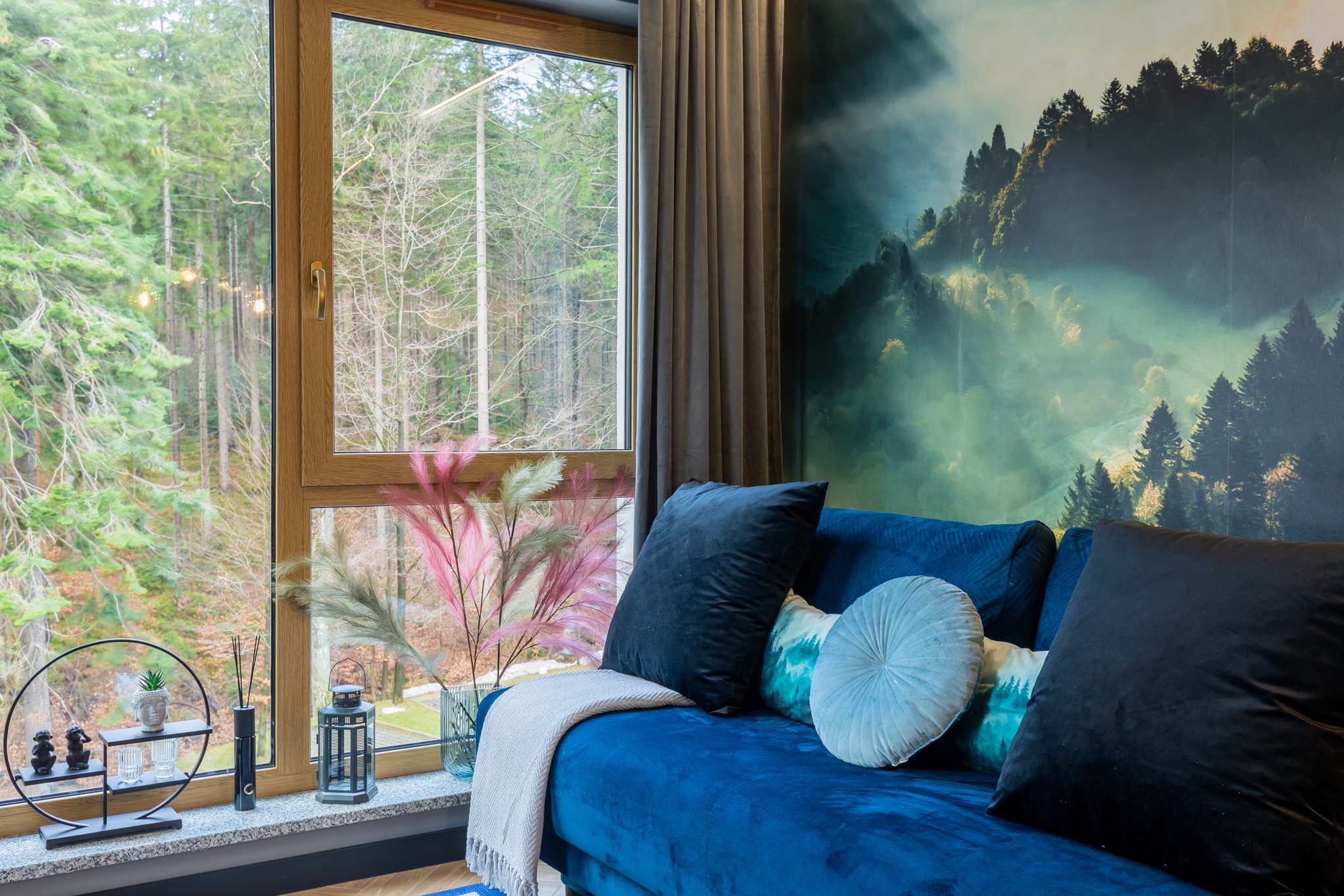 Narożnik obity niebieskim welurem z poduszkami w odcieniach błękitu i czerni na tle okna z widokiem na las i fototapety z motywem górskiej mgły.