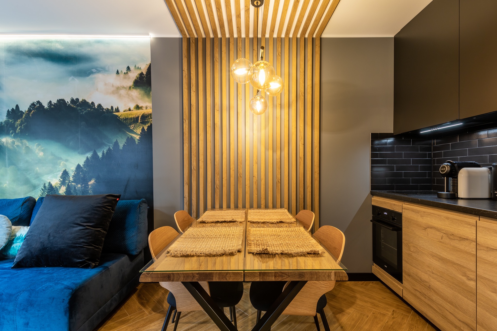 Jadalnia z drewnianym stołem nakrytym czterema słomkowymi podkładkami, oświetlona wiszącą lampą z czterema szklanymi kloszami, z drewnianą boazerią na ścianie i fototapetą z motywem leśnym w tle.