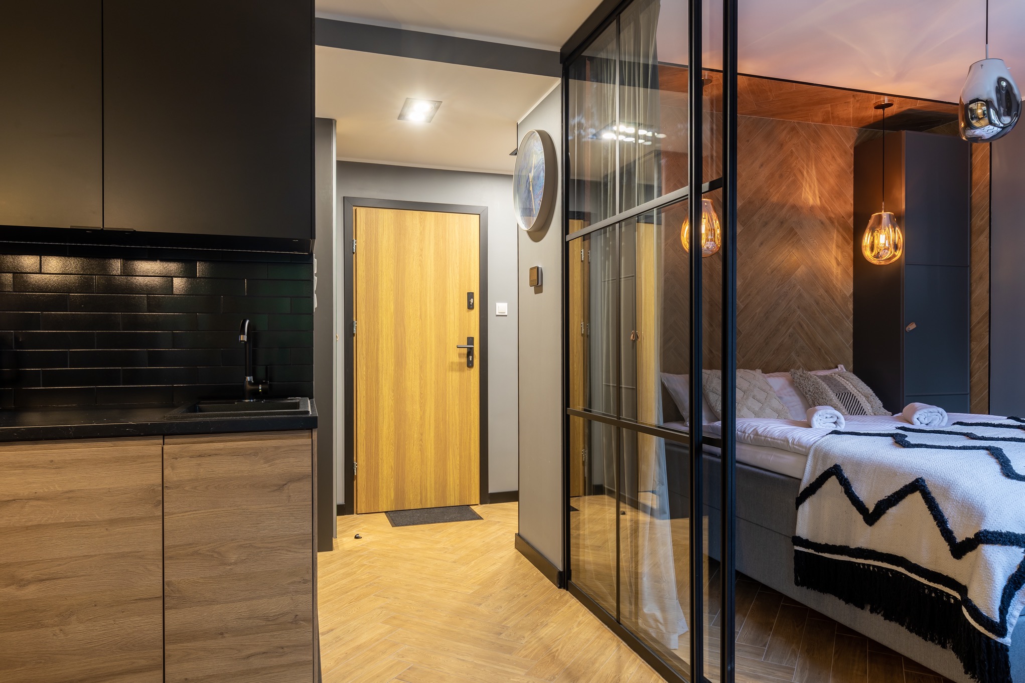 Nowoczesne studio: czarna zabudowa kuchenna z drewnianymi frontami, widok na przedpokój z drewnianymi drzwiami i szklaną ścianą oddzielającą sypialnię z łóżkiem z dekoracyjną narzutą i lampami...