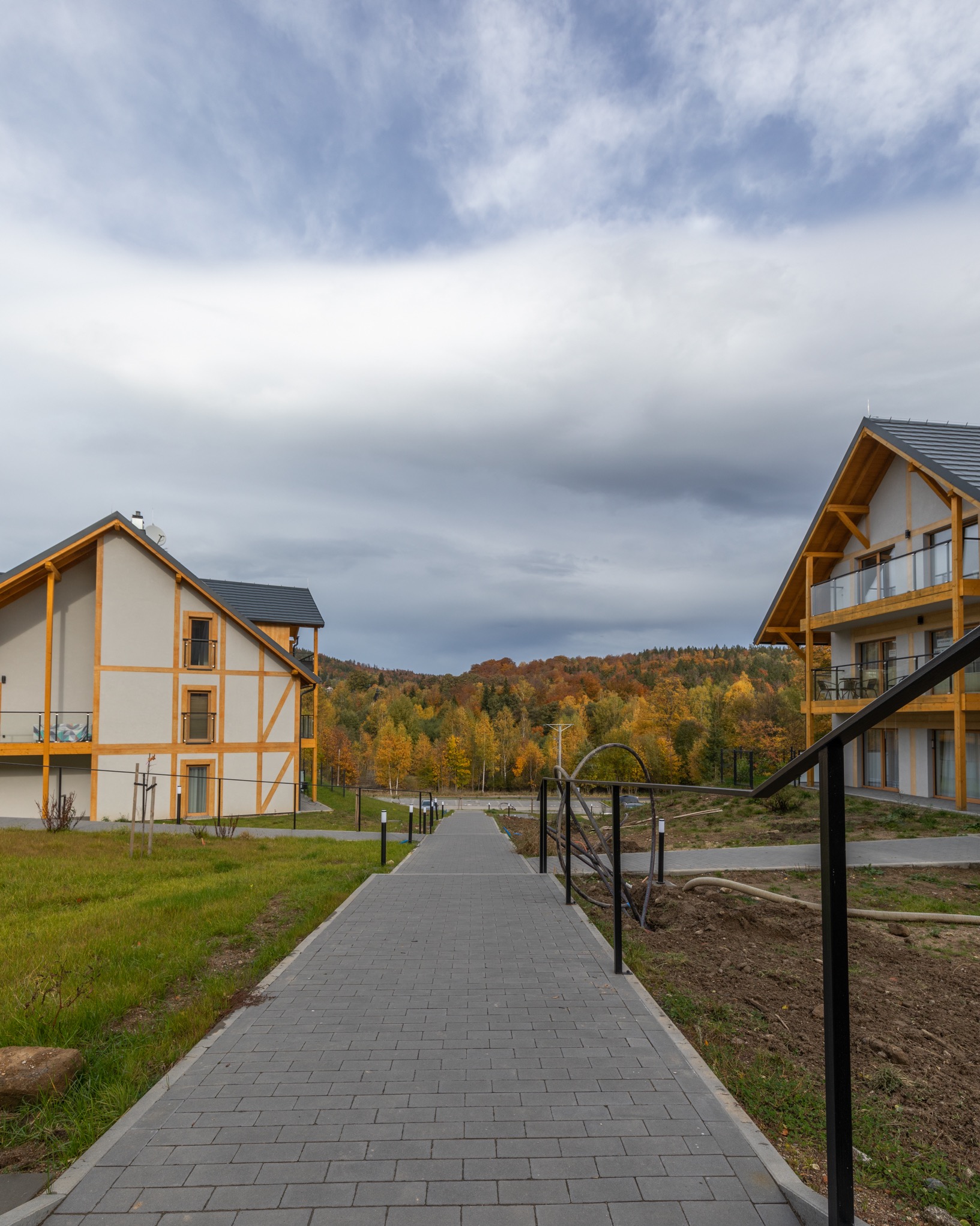 Dwa budynki apartamentowe z drewnianą konstrukcją szkieletową, połączone chodnikiem z kostki brukowej prowadzącym w kierunku lasu w jesiennych barwach, widok z perspektywy chodnika.