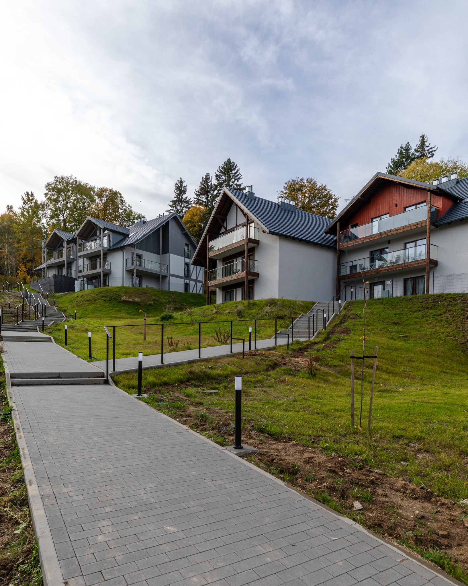 Nowoczesne budynki apartamentowe z balkonami i szklanymi balustradami na wzgórzu, do których prowadzi chodnik z kostki brukowej i schody.