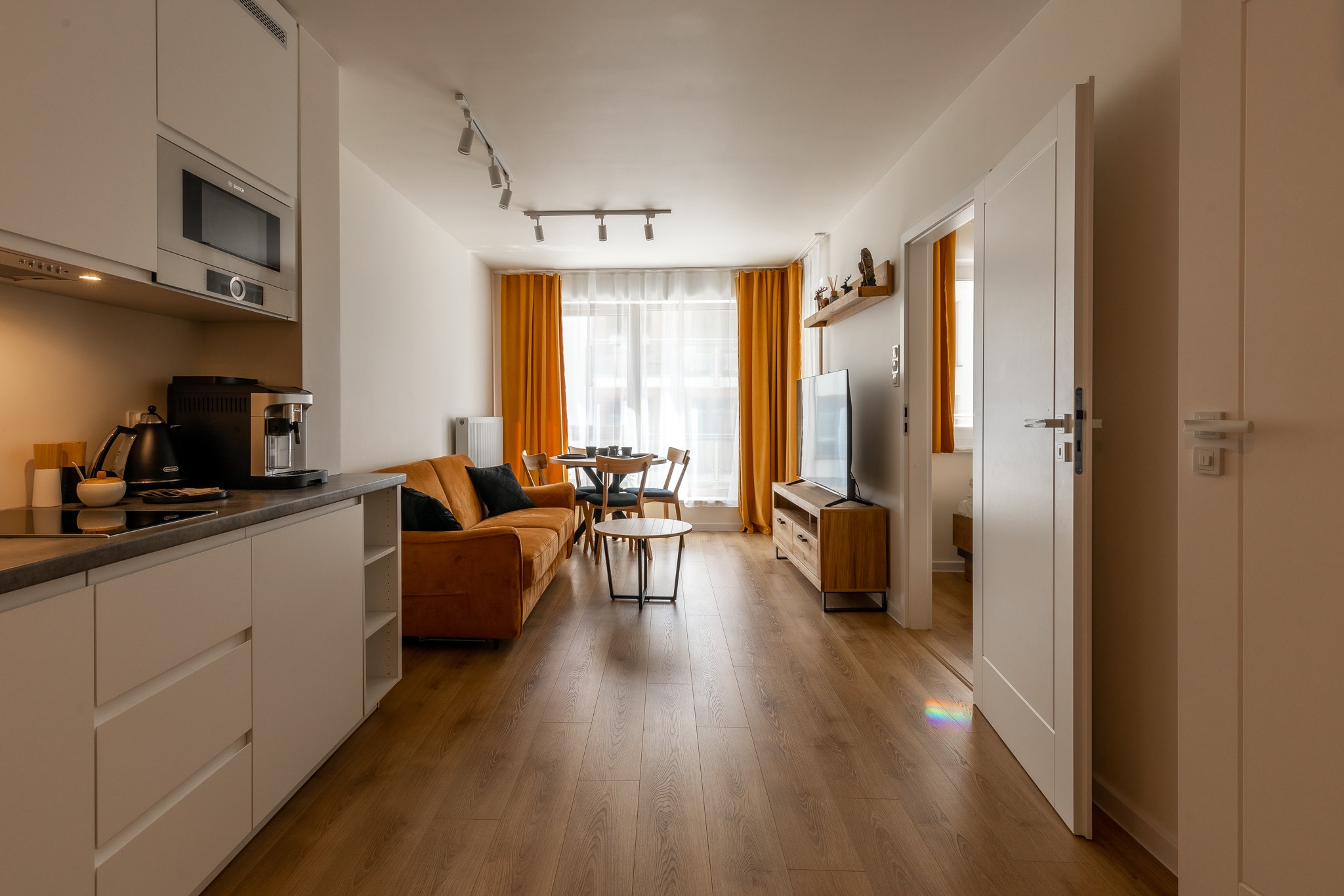 Wnętrze mieszkania z otwartą kuchnią, salonem i jadalnią, z brązową kanapą i pomarańczowymi zasłonami, oświetlone listwą z reflektorami.