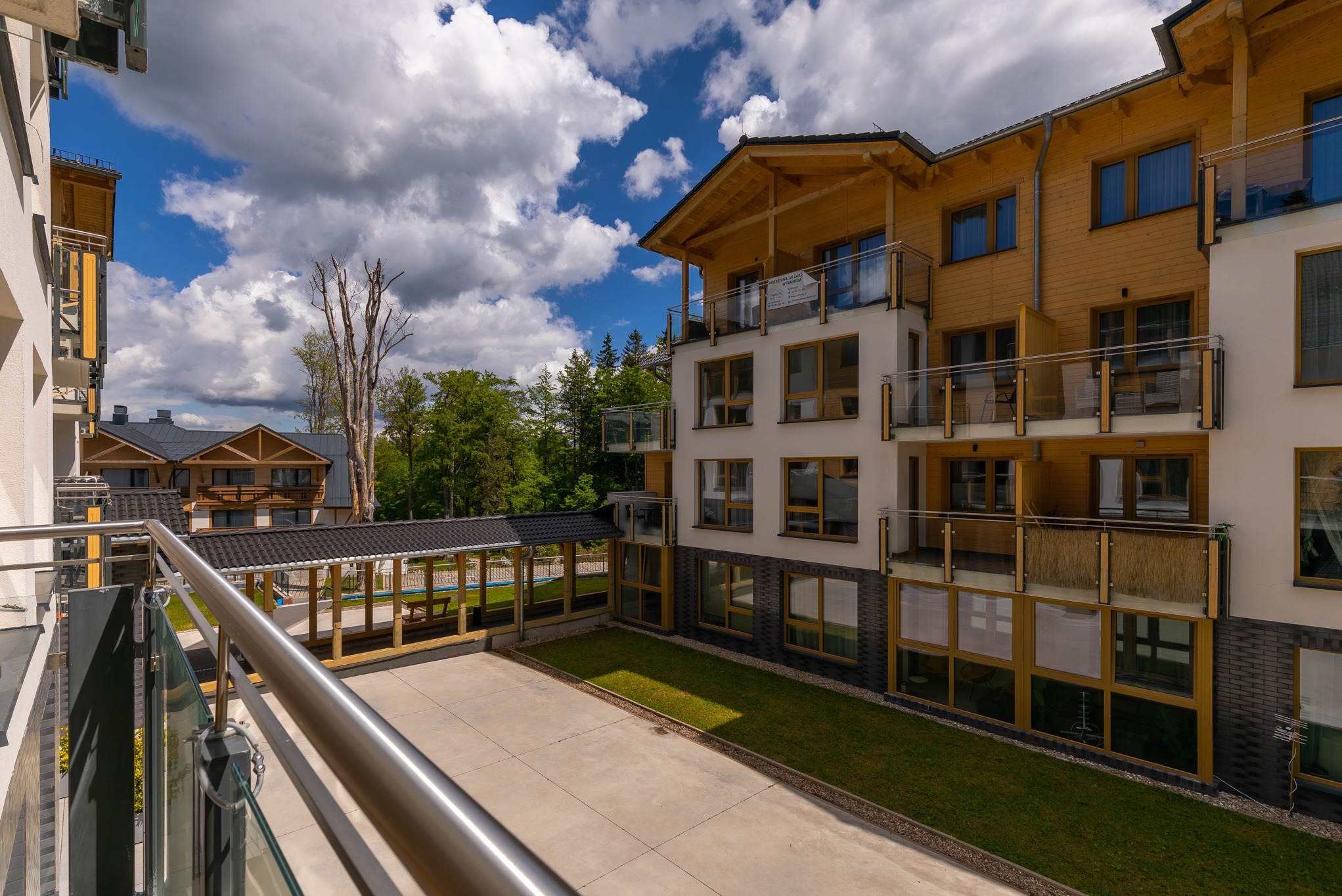 Widok z balkonu na nowoczesny apartamentowiec z drewnianą elewacją, plac z trawnikiem i zadaszoną strefą relaksu z basenem w tle, za budynkiem widoczne drzewa i błękitne niebo z chmurami.