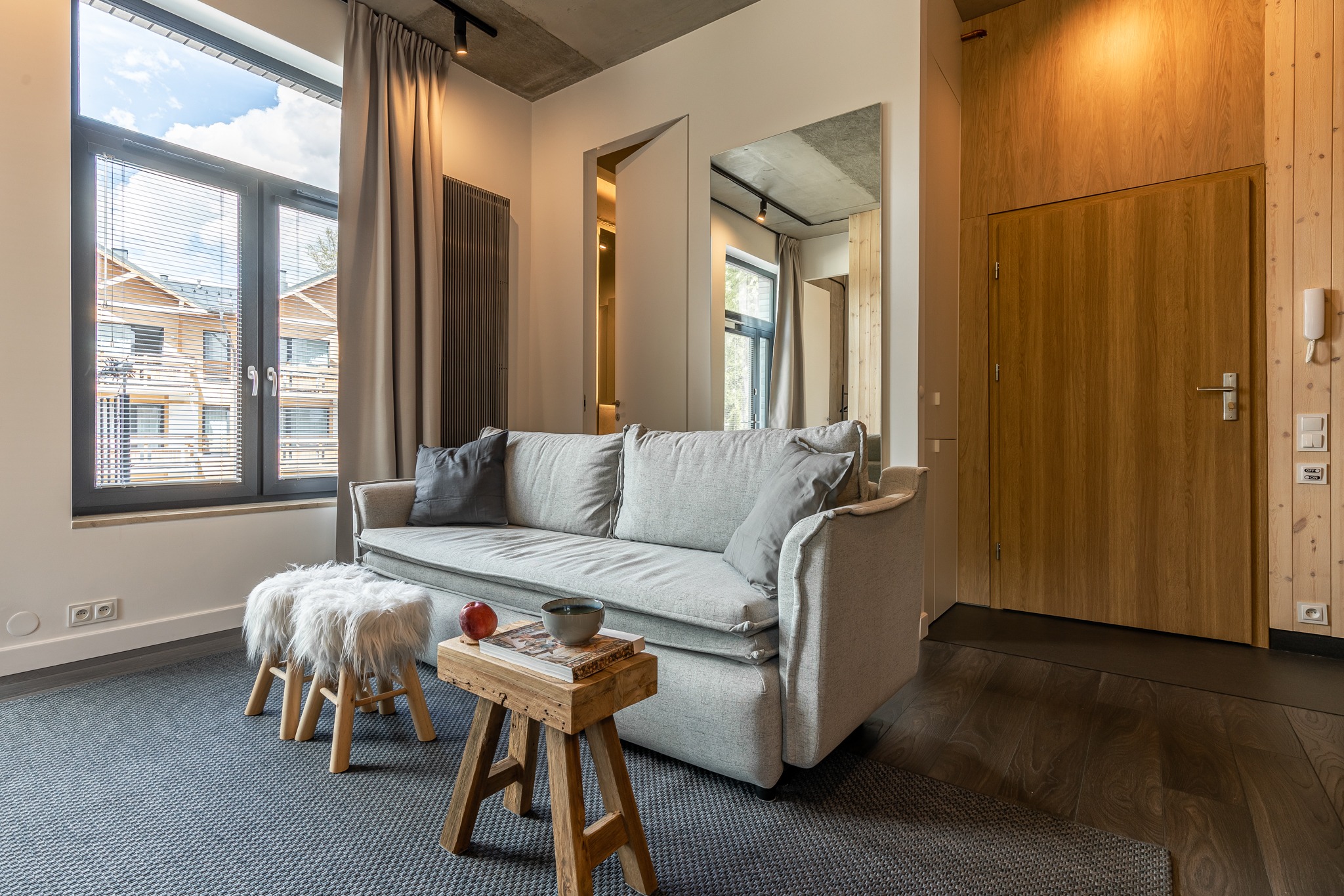 Jasnoszara sofa z poduszkami w nowoczesnym salonie, drewniany stolik z jabłkiem i miseczką, puszysty stołek, okno z widokiem na budynek z czerwonym dachem, drewniana ściana z drzwiami.