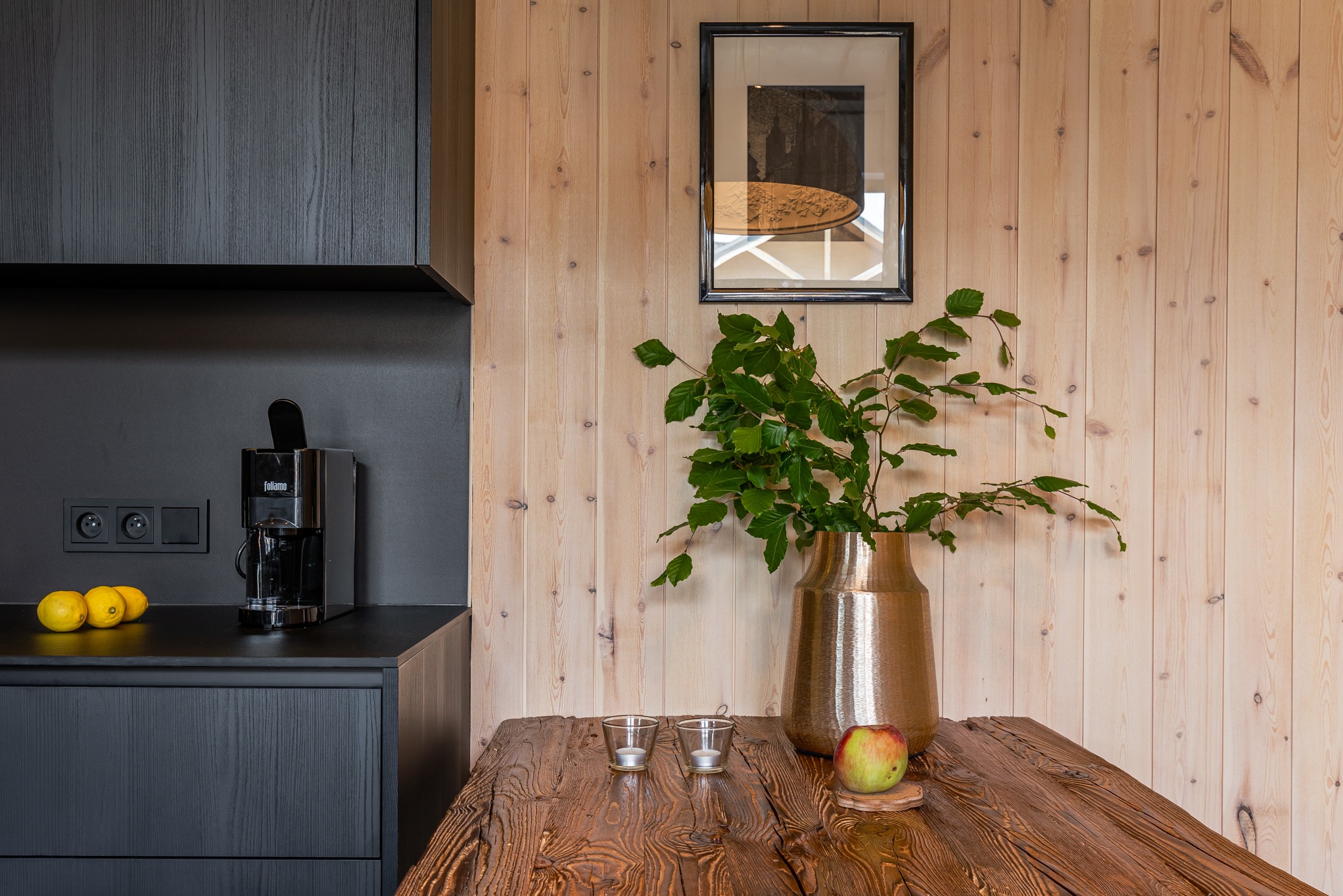 Nowoczesna kuchnia z czarnymi szafkami, drewnianą ścianą i blatem z ciemnego drewna, ozdobiona złotym wazonem z zielonymi gałązkami, jabłkiem, dwoma świecznikami i czarną maszyną do kawy Foliamo.