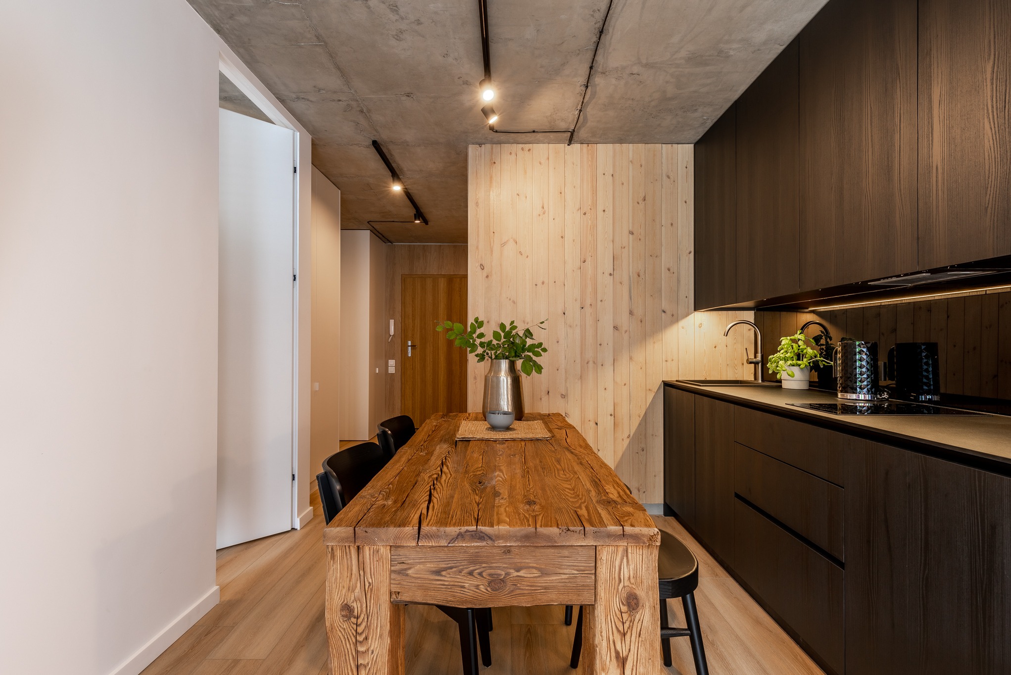 Wnętrze kuchni z drewnianą zabudową, surowym betonowym sufitem i stołem z litego drewna z metalowym wazonem z zielonymi gałązkami.