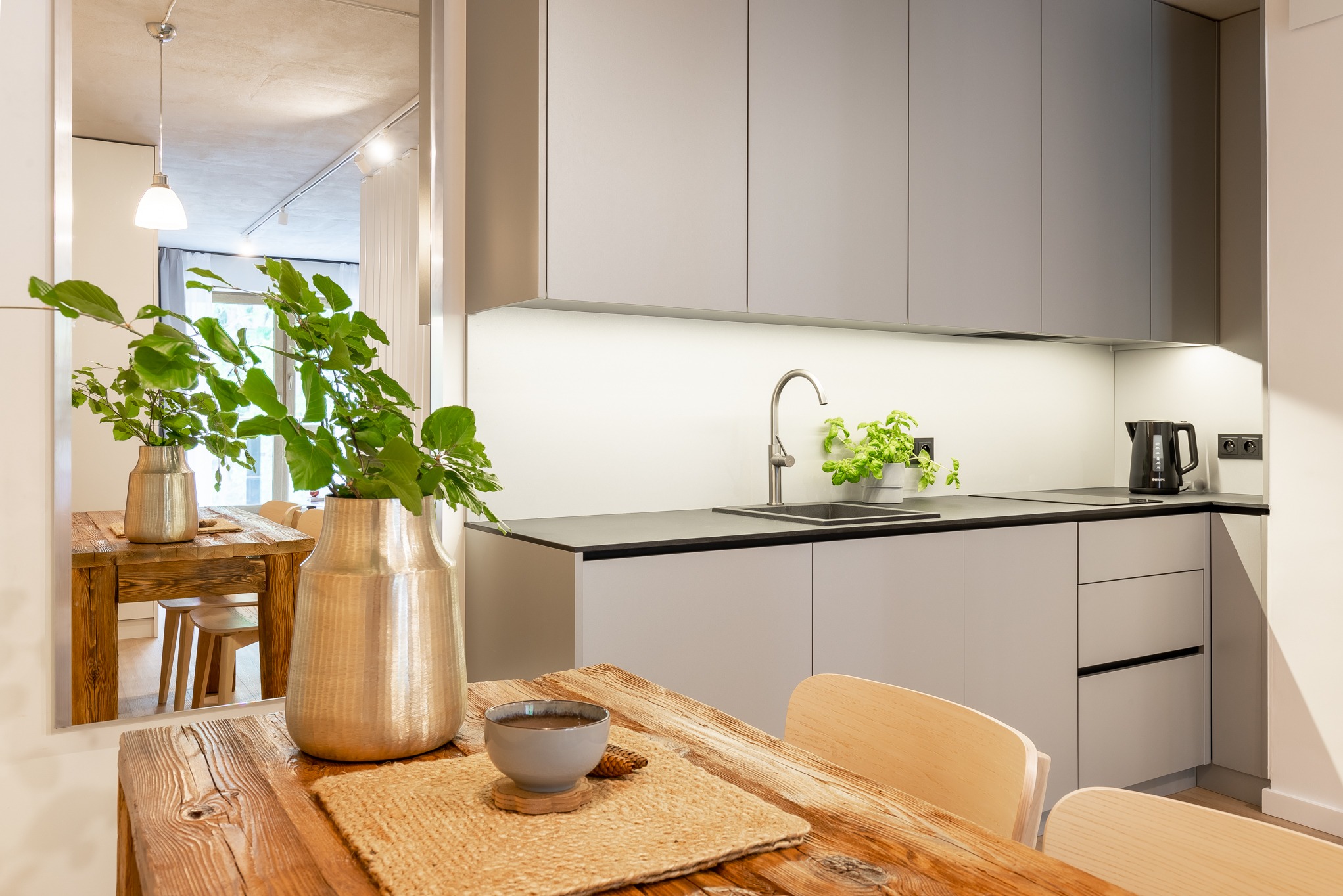 Nowoczesna kuchnia z szarymi szafkami, drewnianym stołem i dekoracyjnymi roślinami w wazonie, odbijająca się w lustrze.