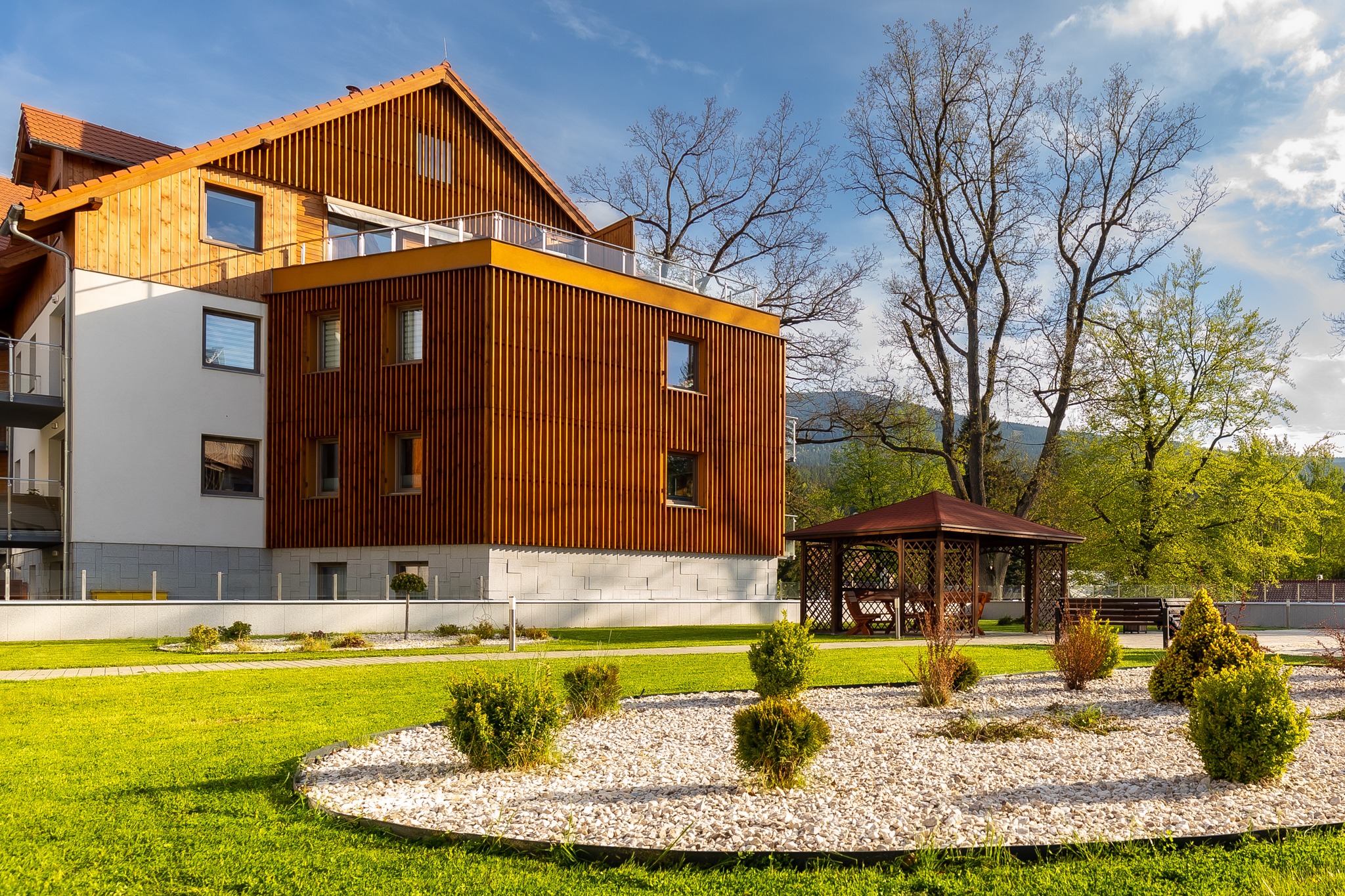 Nowoczesny budynek z drewnianą elewacją i tarasem, otoczony zielonym trawnikiem i ozdobnym klombem z białymi kamieniami oraz altaną w tle.