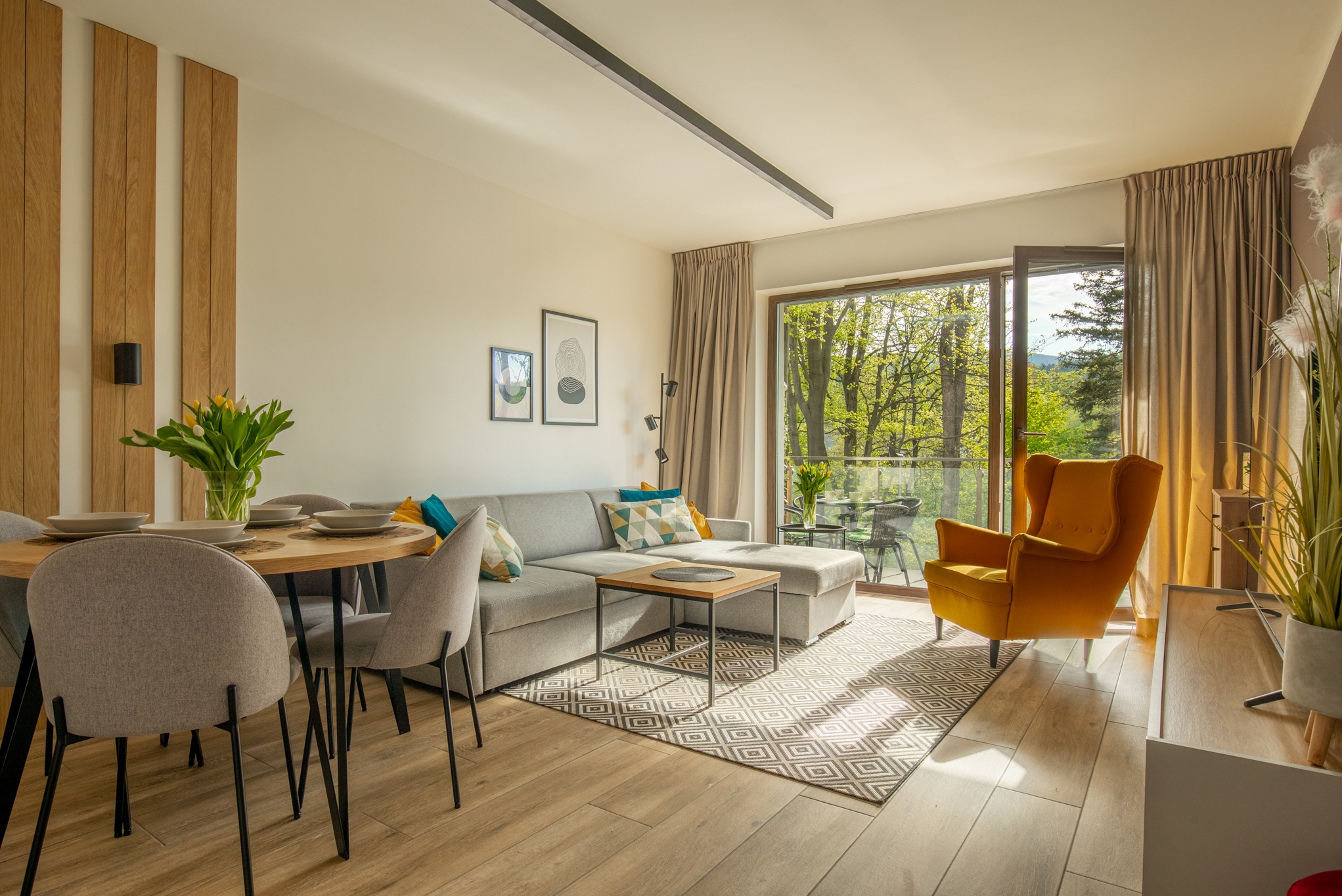 Jasne wnętrze salonu z balkonem, szarą sofą narożną, okrągłym stołem z tulipanami i żółtym fotelem uszakiem na tle zieleni za oknem.