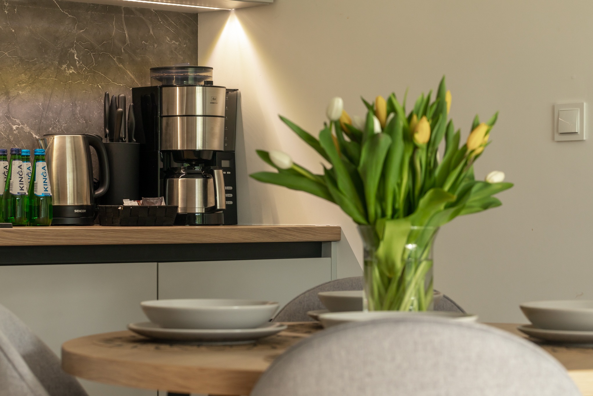 Nowoczesna kuchnia z drewnianym blatem, na którym stoi ekspres do kawy, czajnik, butelki wody i wazon z tulipanami, a na pierwszym planie okrągły stół z nakryciem dla czterech osób.