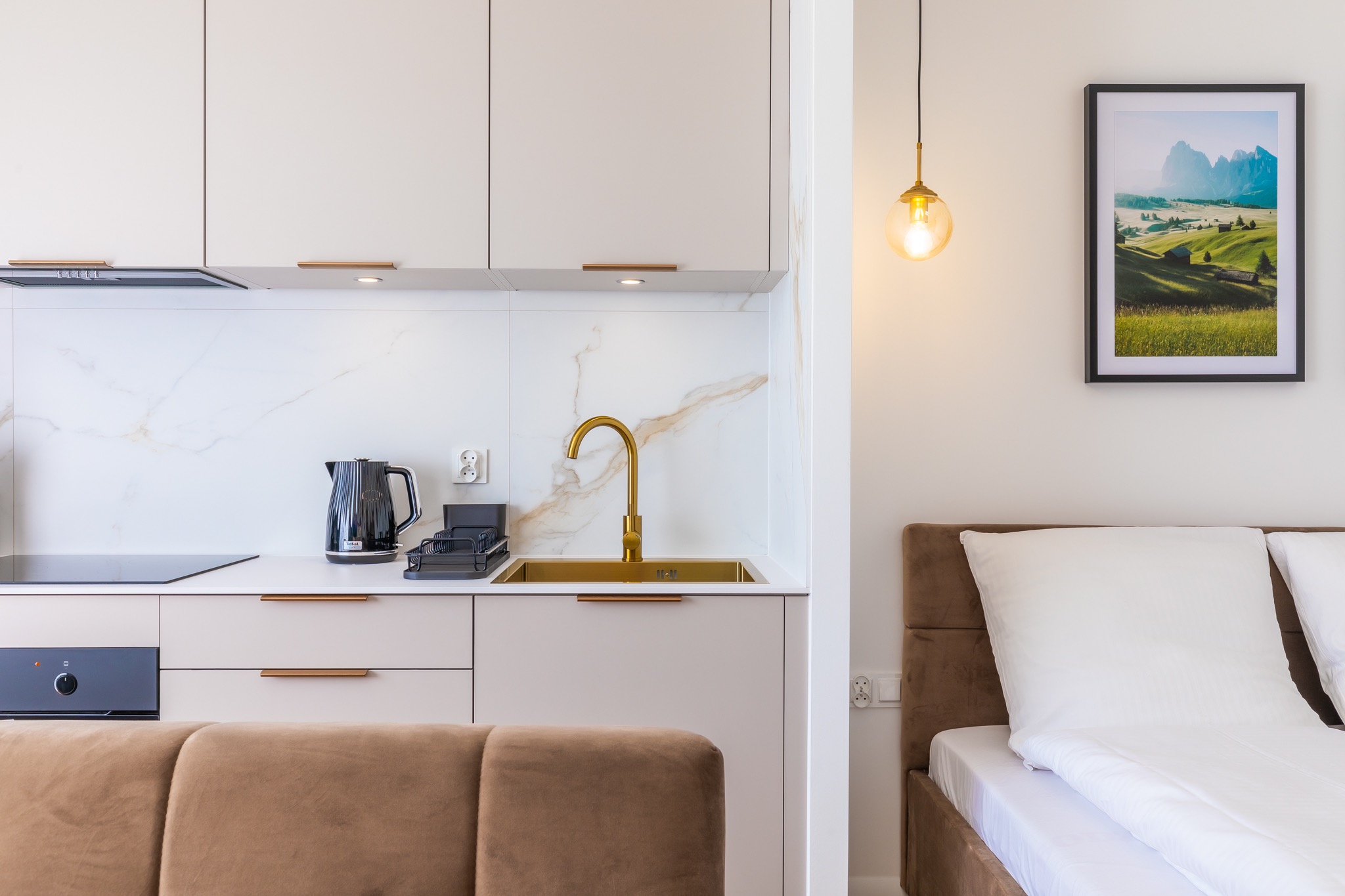 Nowoczesna kuchnia z elementami sypialni: złota armatura, białe szafki, czajnik elektryczny i łóżko z brązowym zagłówkiem w tle.