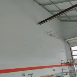 Wnętrze hali produkcyjnej po świeżym pomalowaniu ścian na biało, widoczne metalowe elementy konstrukcyjne pod sufitem, częściowo otwarte drzwi garażowe i rusztowanie przy ścianie.