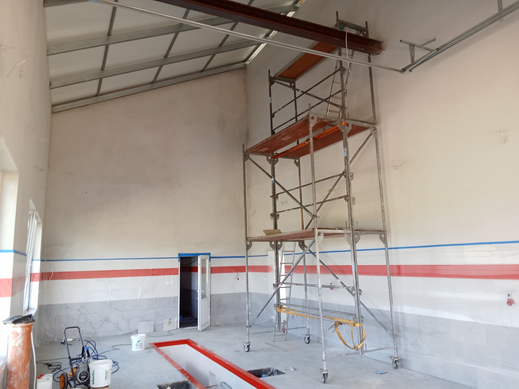 Wnętrze warsztatu samochodowego w trakcie malowania: widoczne rusztowanie, agregat malarski, zabezpieczone folią ściany z czerwonym pasem i szarym dołem, biały sufit z metalową konstrukcją.