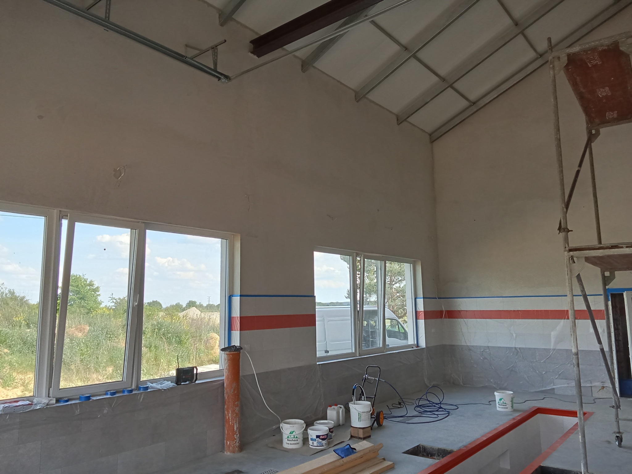 Wnętrze budynku w trakcie prac wykończeniowych: ściany z pomalowanym pasem, okna, sprzęt malarski, rusztowanie i materiały budowlane.