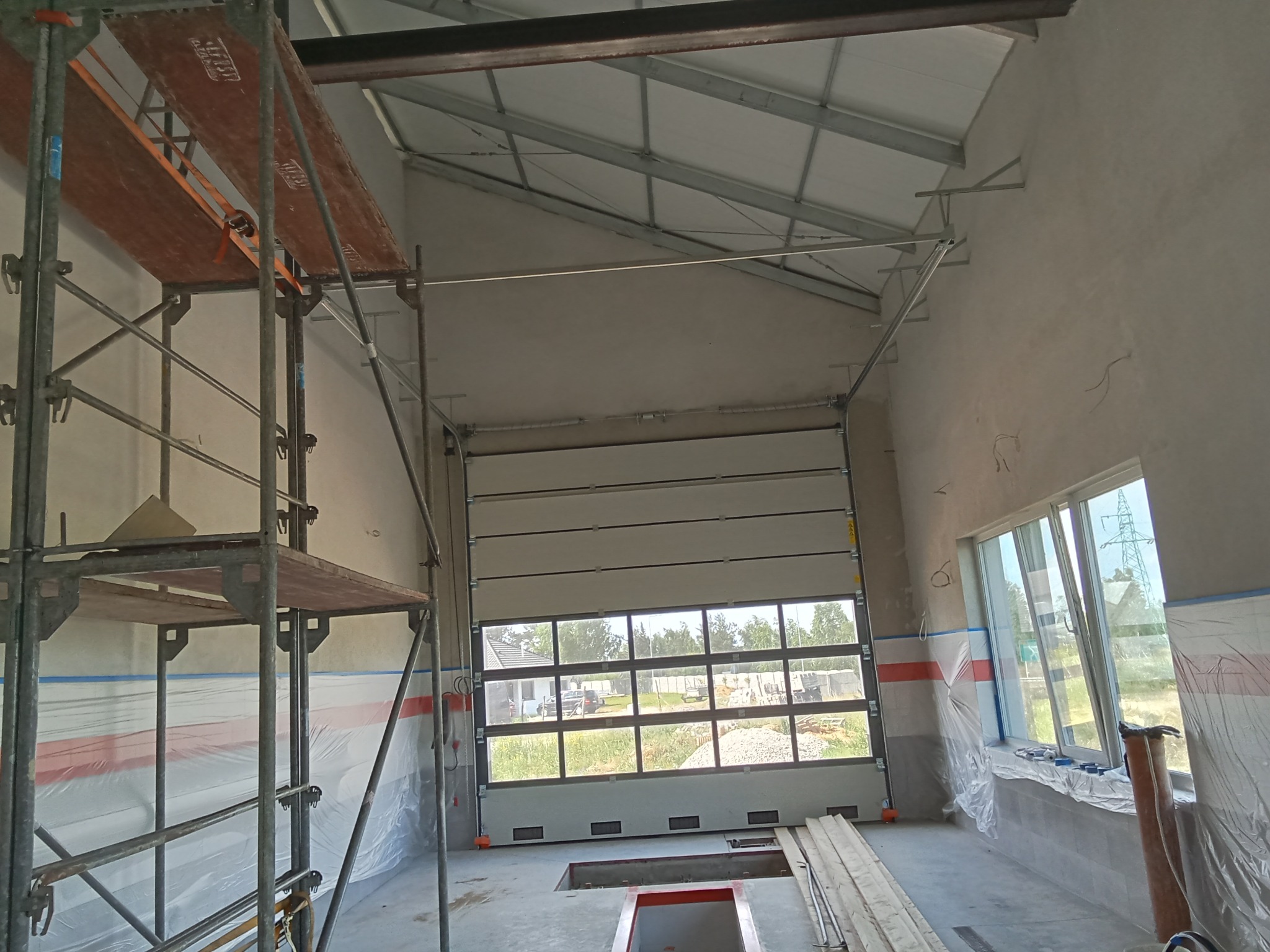 Wnętrze hali w trakcie prac wykończeniowych, widoczne rusztowanie, częściowo zakryte folią okna i brama garażowa z przeszkleniami, kanał samochodowy w podłodze.