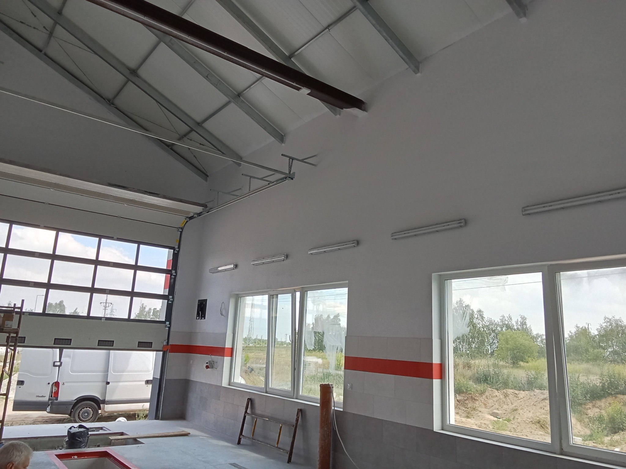 Wnętrze hali warsztatowej po malowaniu, widoczne pomalowane ściany na biało, okna z widokiem na zewnątrz, częściowo otwarta brama garażowa i biały bus na zewnątrz.