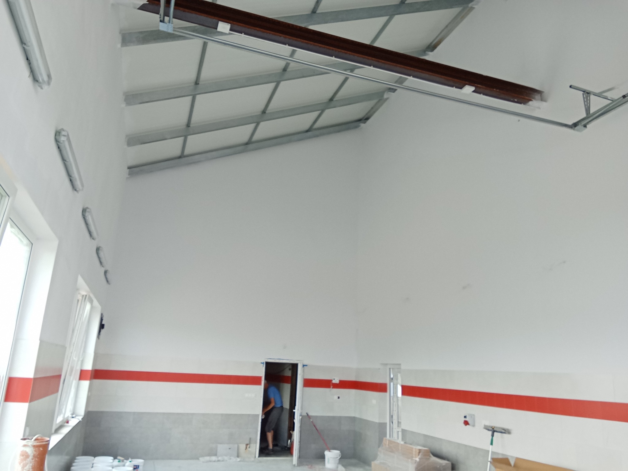 Wnętrze hali po malowaniu, widoczny fragment sufitu z metalową konstrukcją, białe ściany z poziomym czerwonym pasem, okna i drzwi, oraz puste pojemniki po farbach.