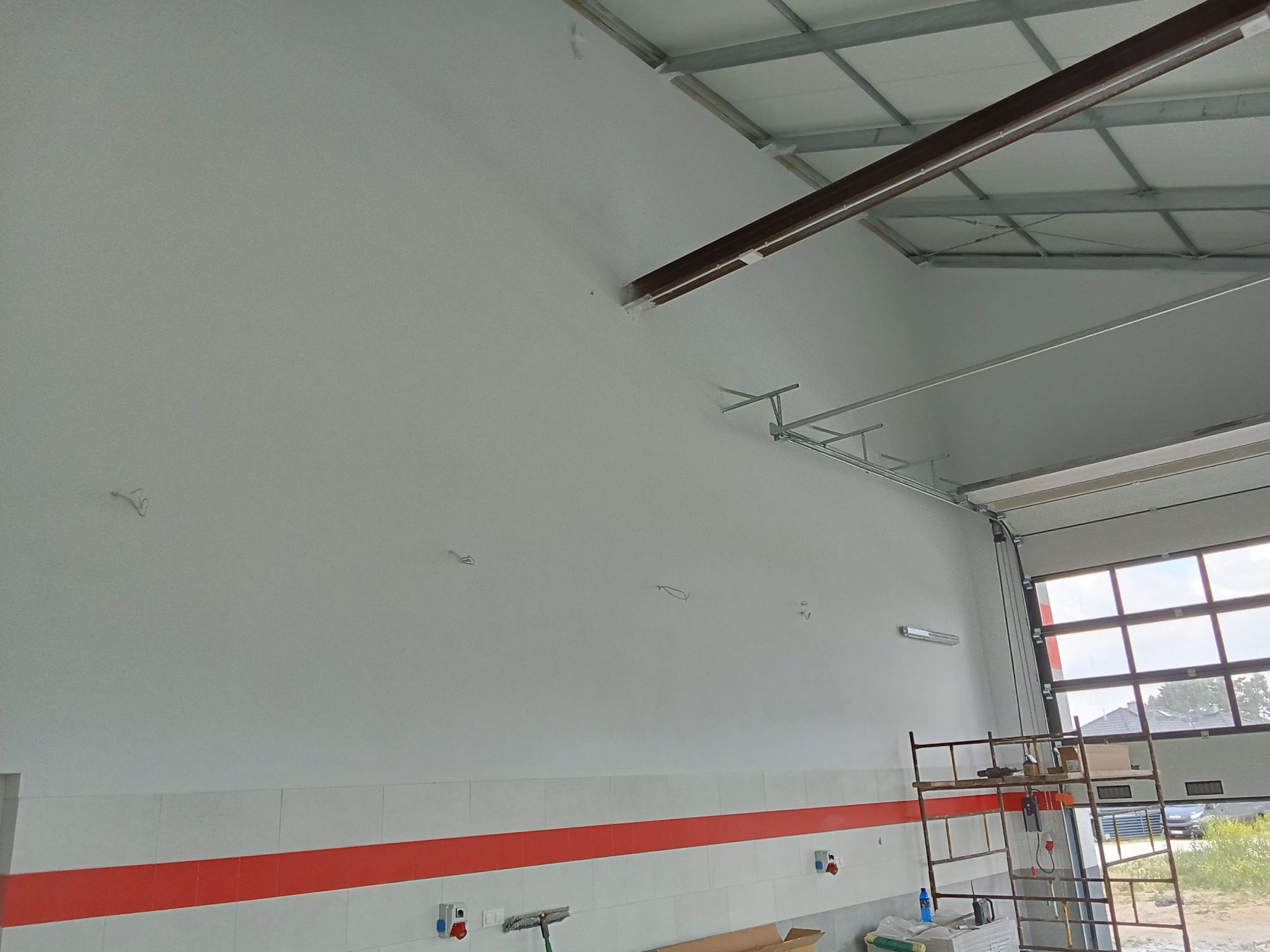 Wnętrze hali produkcyjnej po świeżym pomalowaniu ścian na biało, widoczne metalowe elementy konstrukcyjne pod sufitem, częściowo otwarte drzwi garażowe i rusztowanie przy ścianie.