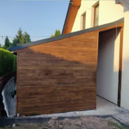 Jednospadowa wiata samochodowa z szopą narzędziową przylegająca do ściany domu, wykonana z ciemnego drewna, z widocznym wejściem do szopy i fragmentem elewacji budynku.