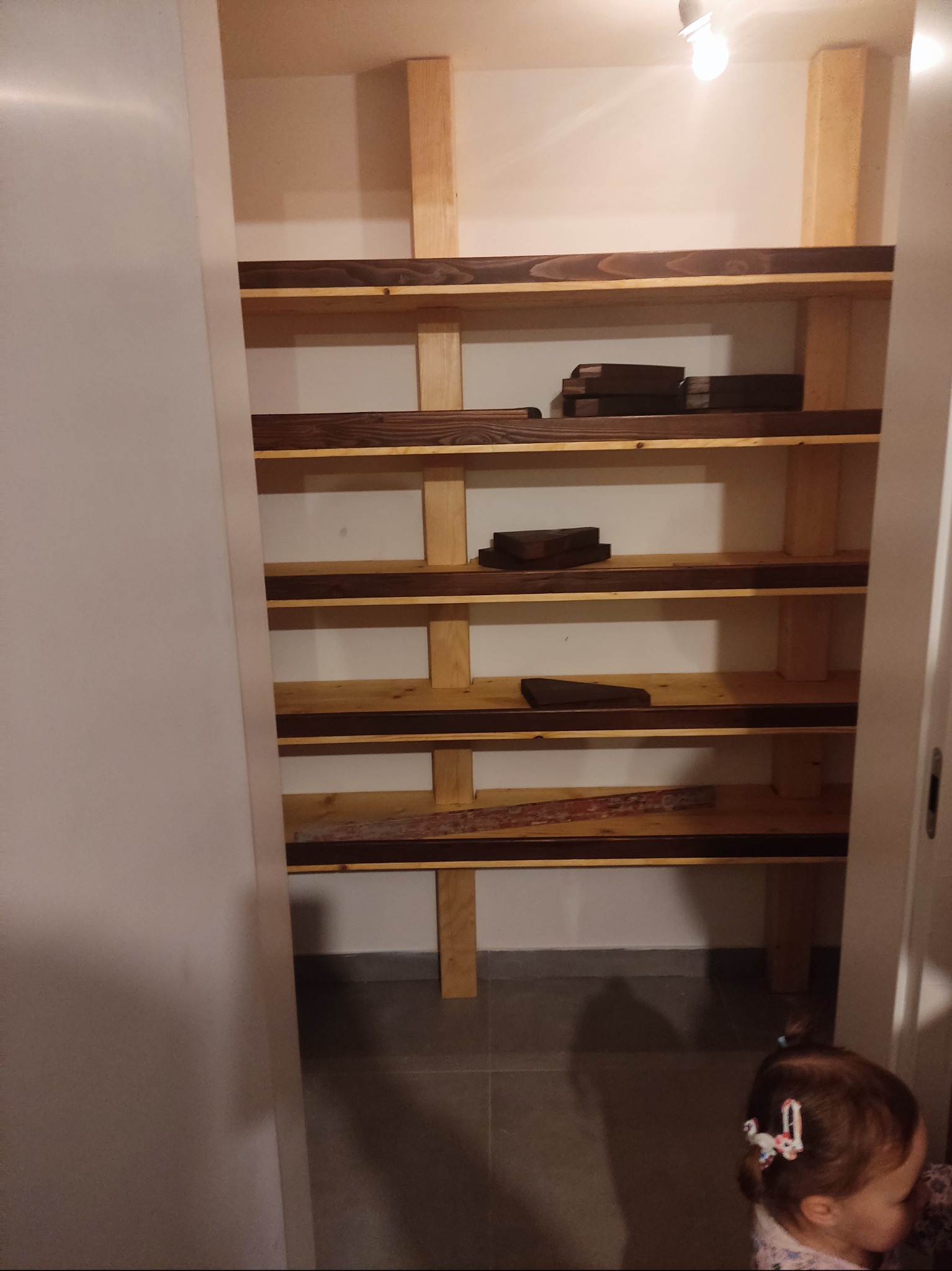 Wnętrze spiżarni z drewnianymi półkami na konstrukcji z jasnego drewna, wykończonymi ciemniejszym, olejowanym drewnem, widoczne deski i fragmenty drewna na półkach, u góry żarówka, w dolnej części...