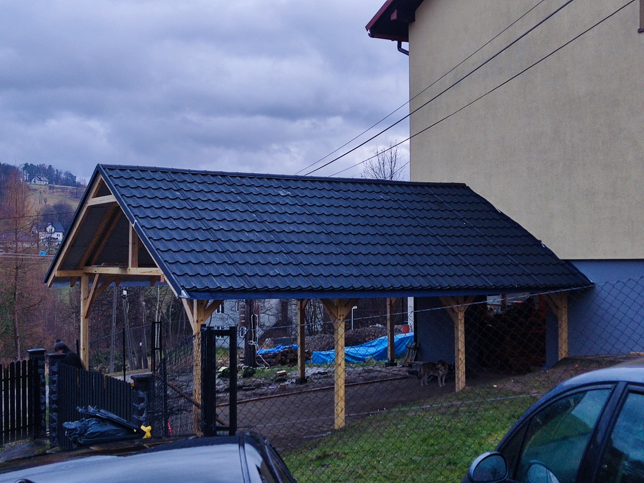 Dwuspadowa wiata drewniana z ciemną dachówką, przylegająca do ściany domu, widoczna zza ogrodzenia, z ułożonym drewnem wewnątrz i psem w pobliżu.