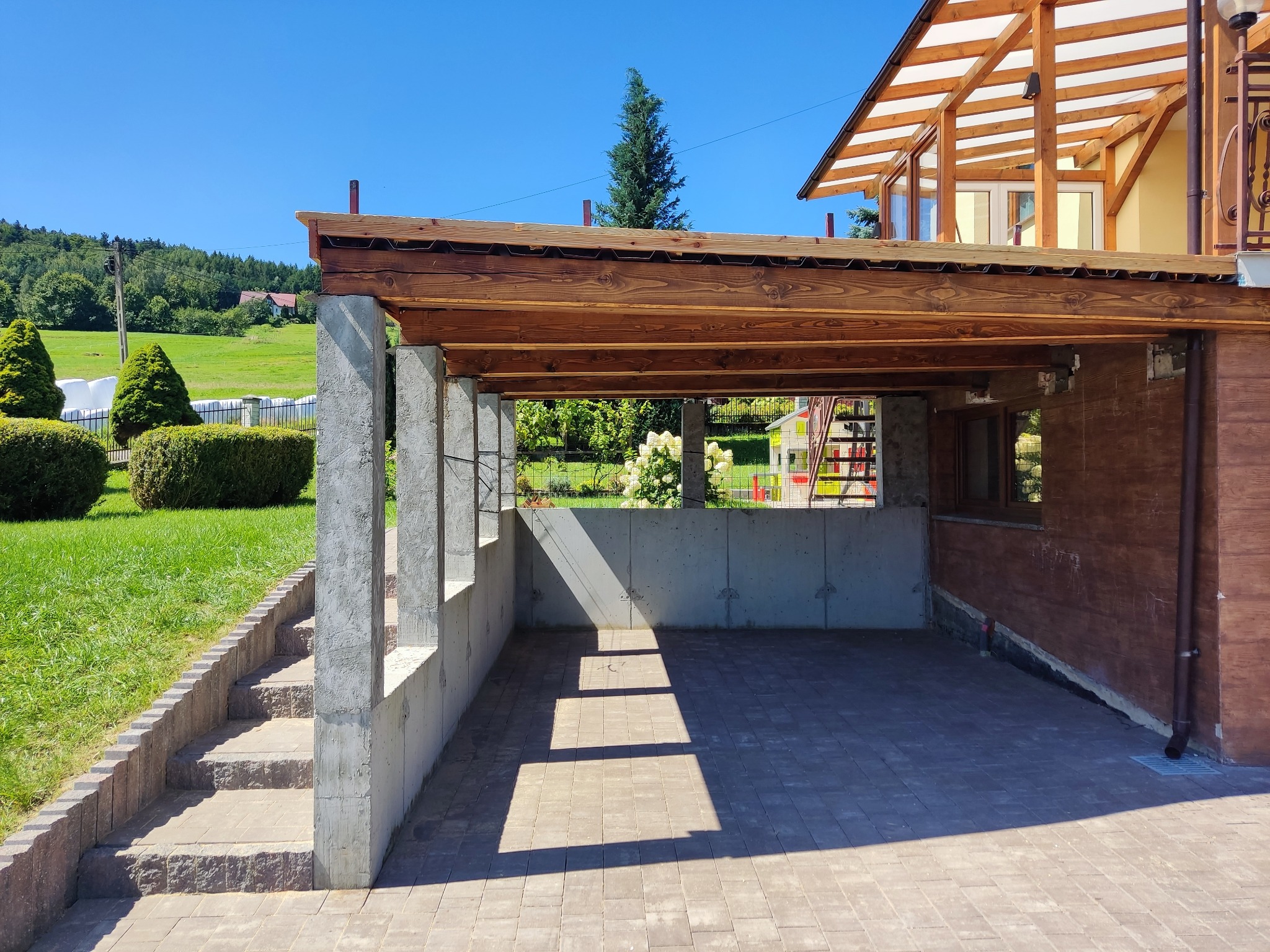Zadaszenie tarasu z drewnianą konstrukcją wspartą na betonowych słupach i blachą trapezową, widok z zewnątrz, z fragmentem schodów wejściowych i trawnika.